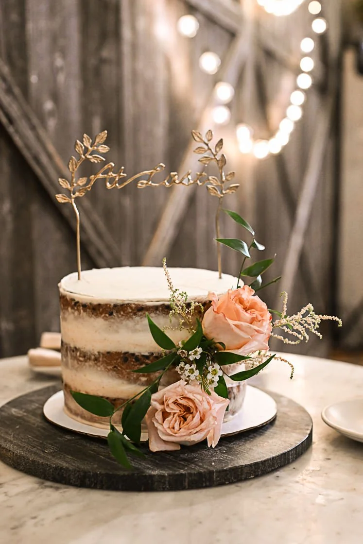 A rustic, semi-naked birthday cake decorated with light pink roses, greenery, and white baby's breath, topped with a gold 'birthday' cake topper, on a wooden cake stand with string lights in the background.