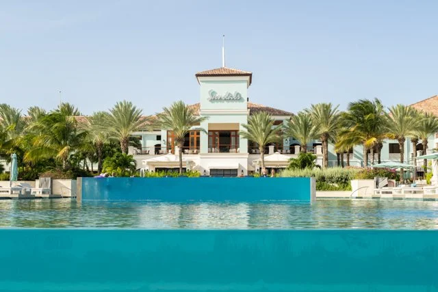 View of a luxurious resort with a large swimming pool, palm trees, and a building with a tower in the background under a clear blue sky.
