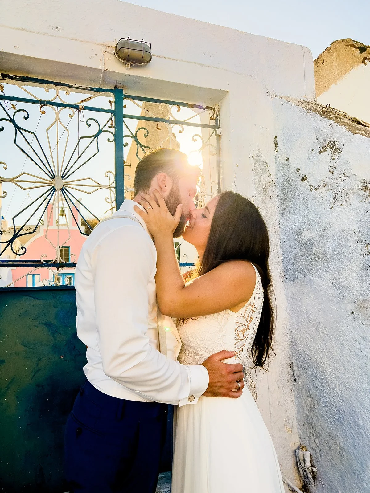 A romantic moment between a man and a woman sharing a kiss outdoors at sunset, with the sun behind them and a white wall and colorful building in the background.