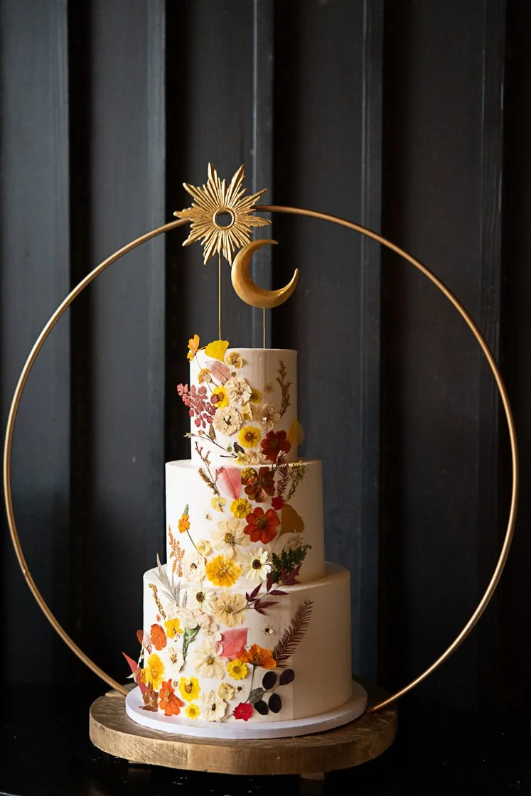 Elegant three-tier wedding cake decorated with colorful edible flowers, displayed on a wooden stand with a decorative gold circle frame featuring a sun and crescent moon embellishments.