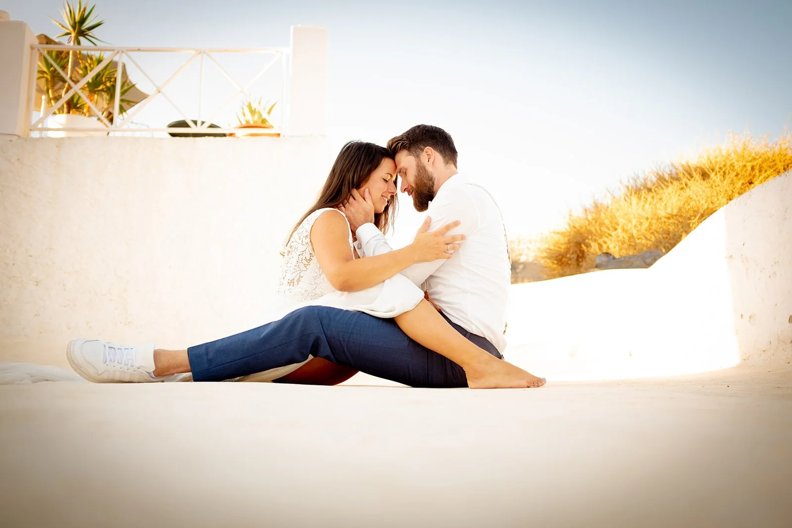 A couple sitting on the ground in a romantic embrace, touching foreheads with eyes closed, outdoors with white walls and blue sky.