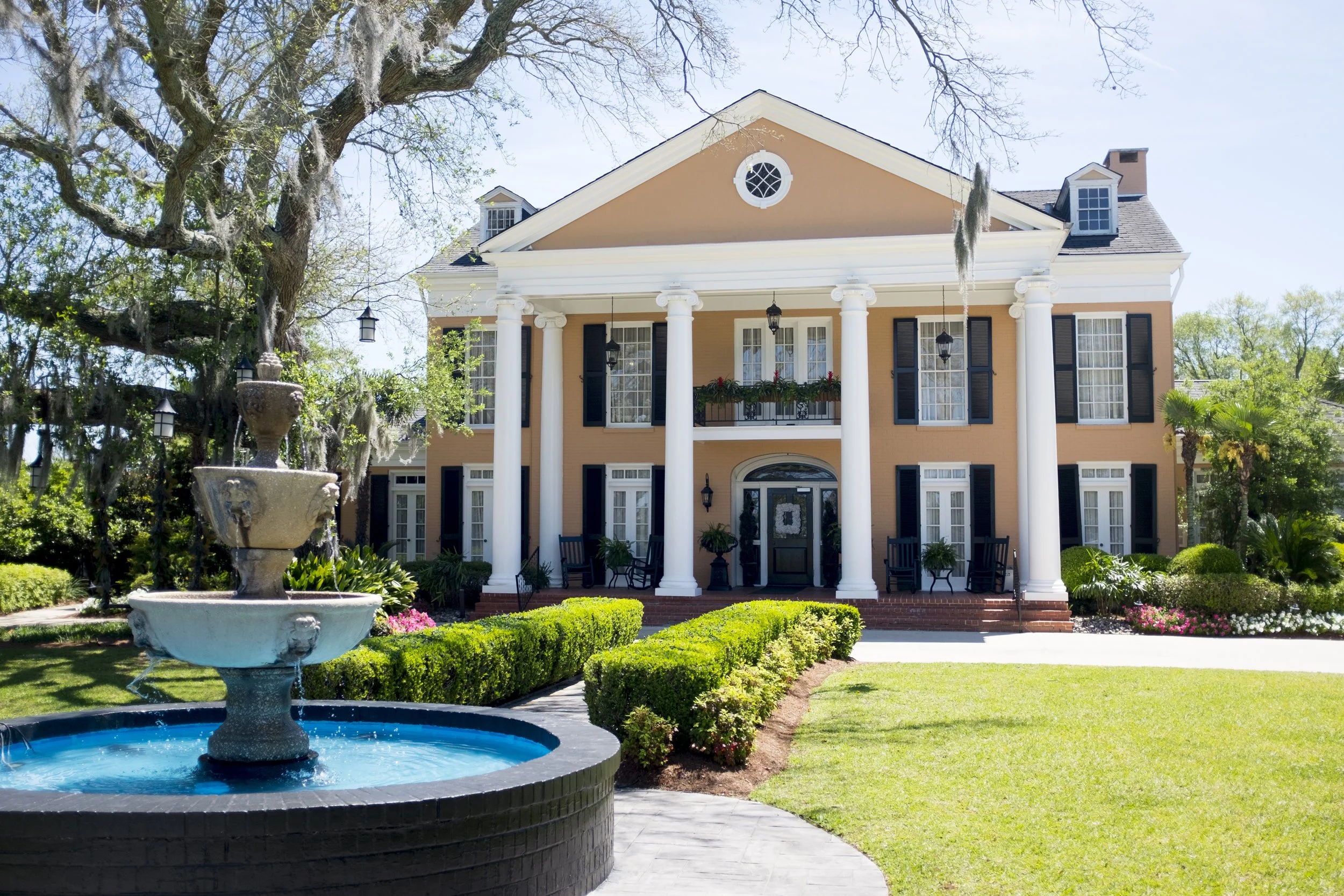 Front view of a large peach-colored house with white columns, black shutters, and a gabled roof, surrounded by a well-maintained garden with a fountain in the foreground and large trees.