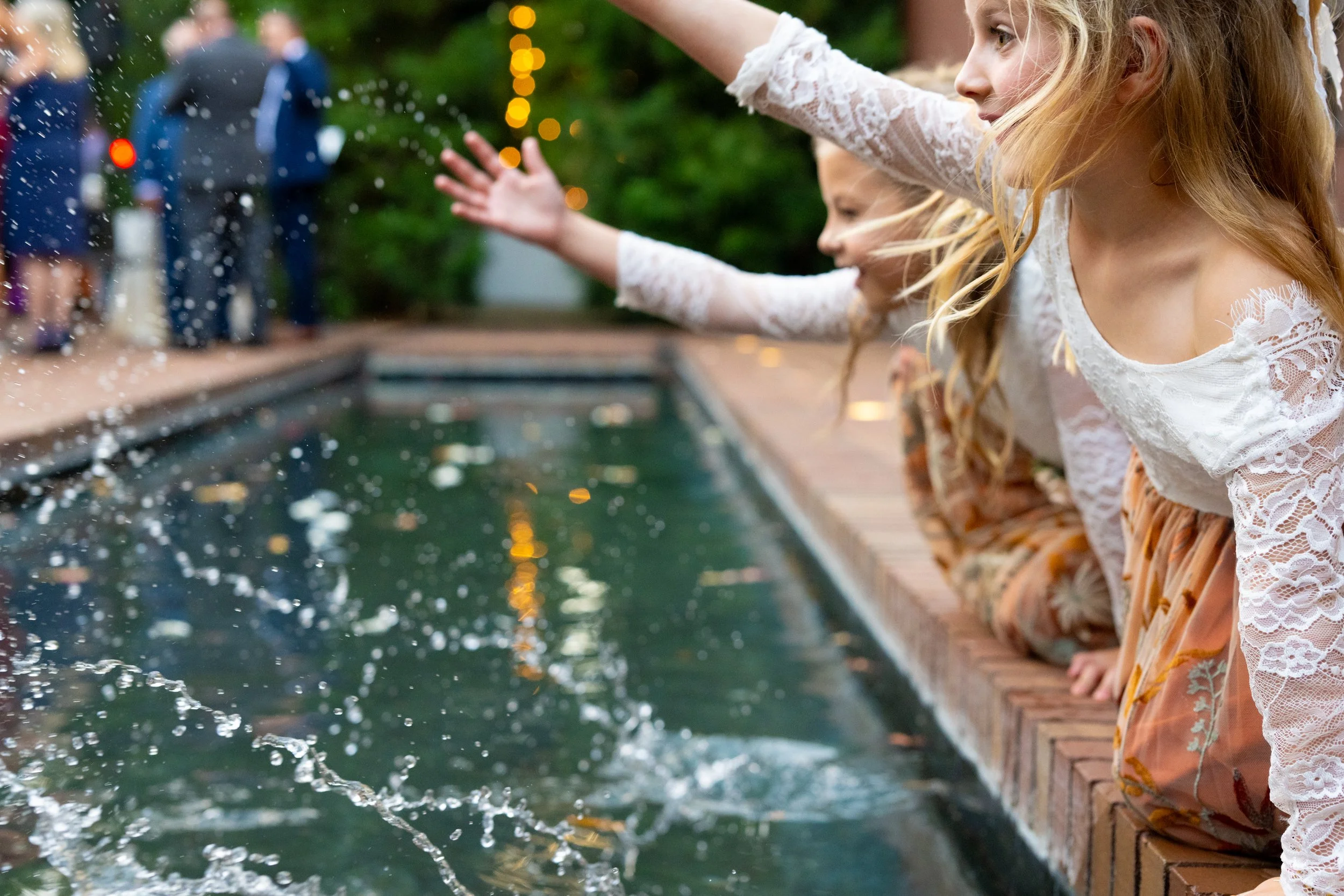 Young girls with blonde hair wearing white lace tops and orange skirts sitting on a brick ledge, reaching out to touch water in a pool, with blurred party guests in the background.