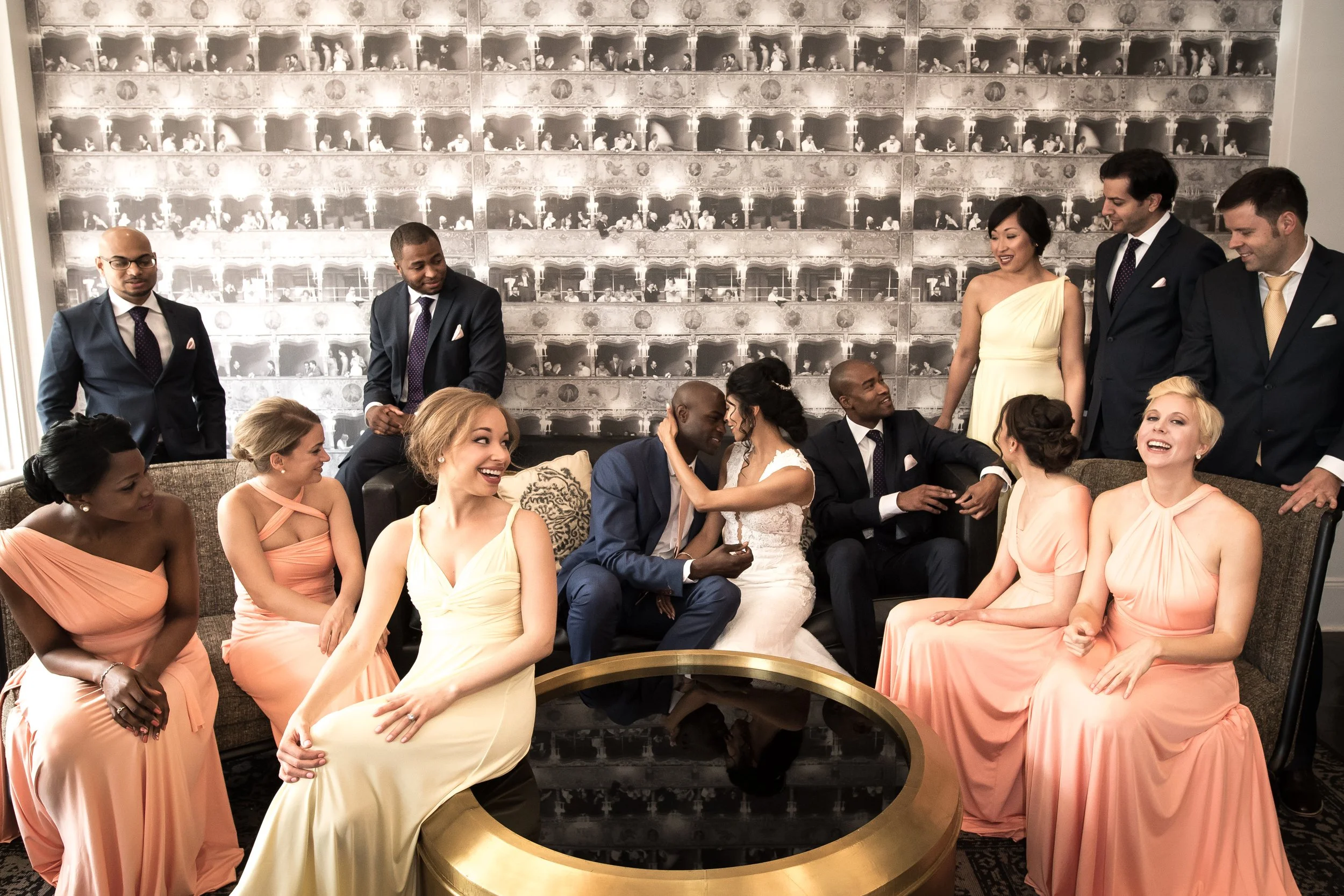 A diverse group of people dressed in formal attire, gathered in a decorated room with a large wall mural resembling theater boxes filled with people, smiling and engaging with each other, possibly celebrating a special occasion or attending a formal 