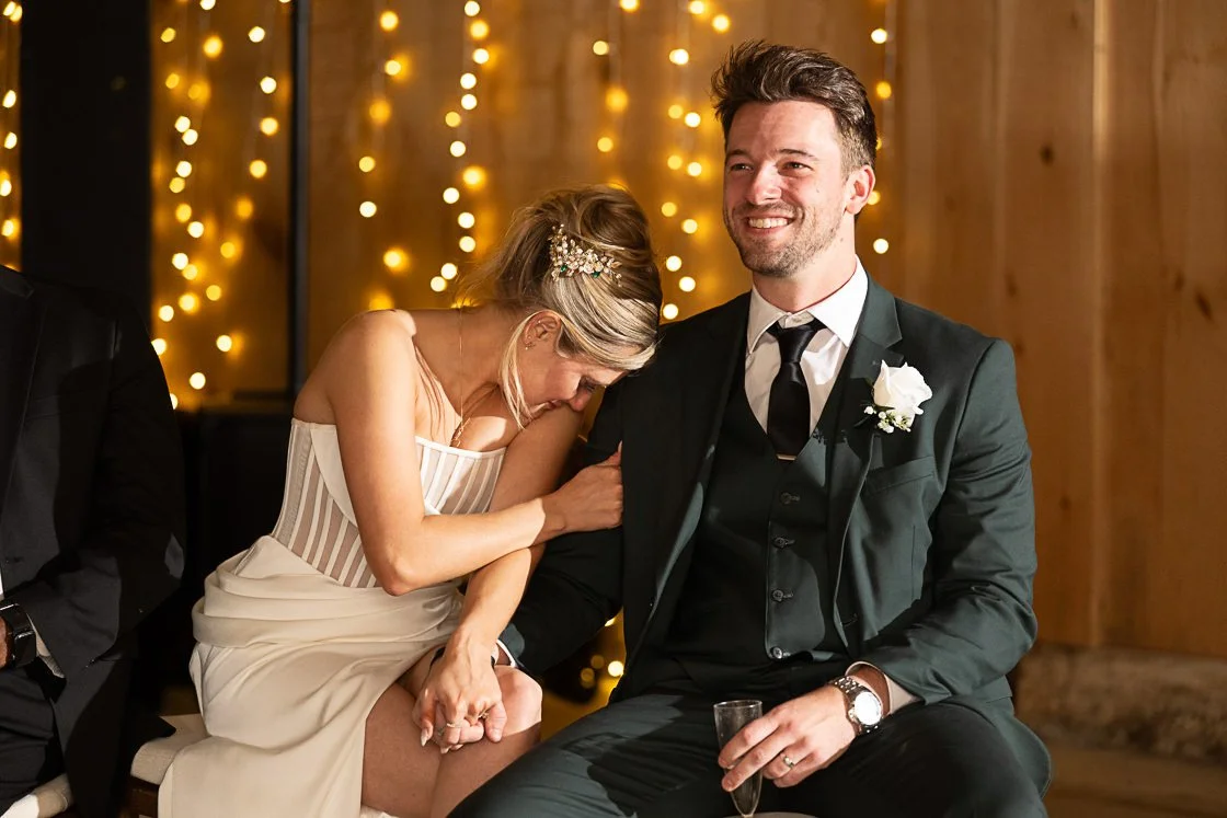 A bride leaning on a groom's shoulder, both smiling, during a wedding reception with warm fairy lights in the background.