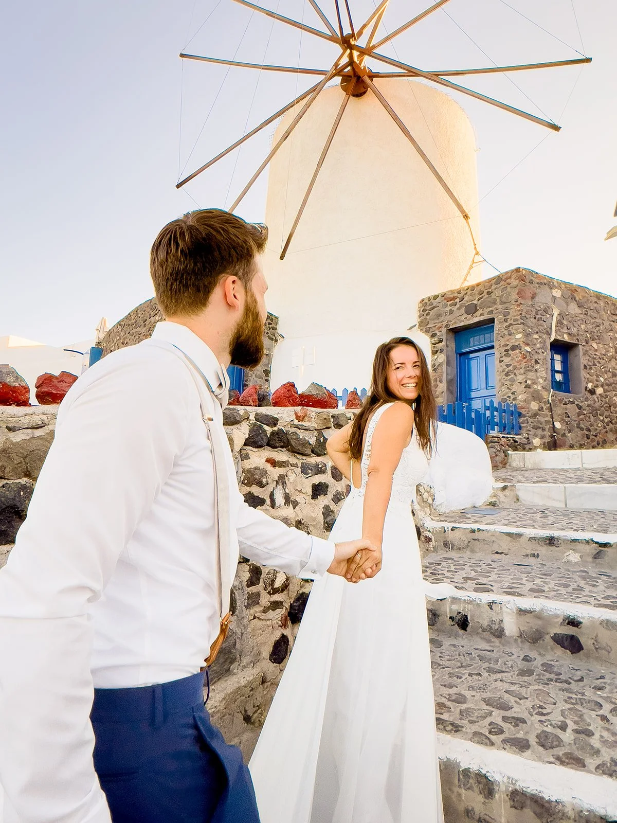 A young couple holding hands and smiling, walking up stone steps near a traditional windmill with a white base and wooden blades, in a scenic location with stone buildings and a cobblestone path.