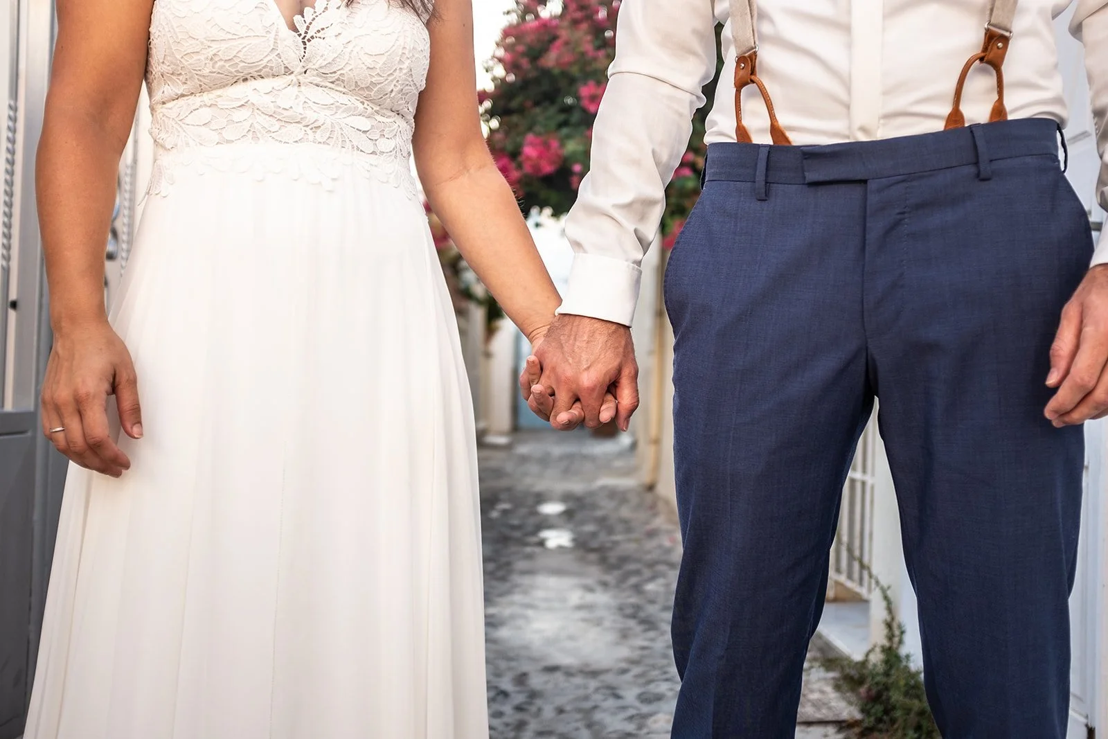 A bride and groom holding hands during their wedding ceremony. The bride is wearing a white lace dress, and the groom is wearing dark blue pants with suspenders and a white shirt.