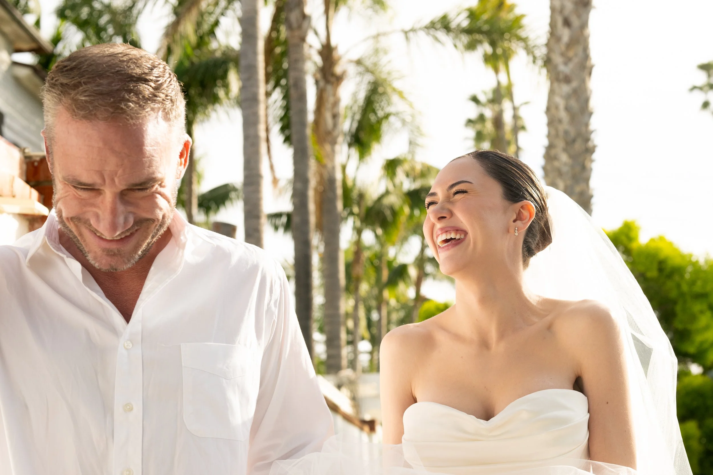 A couple, a man and a woman, outdoors with palm trees in the background, smiling and looking joyful.