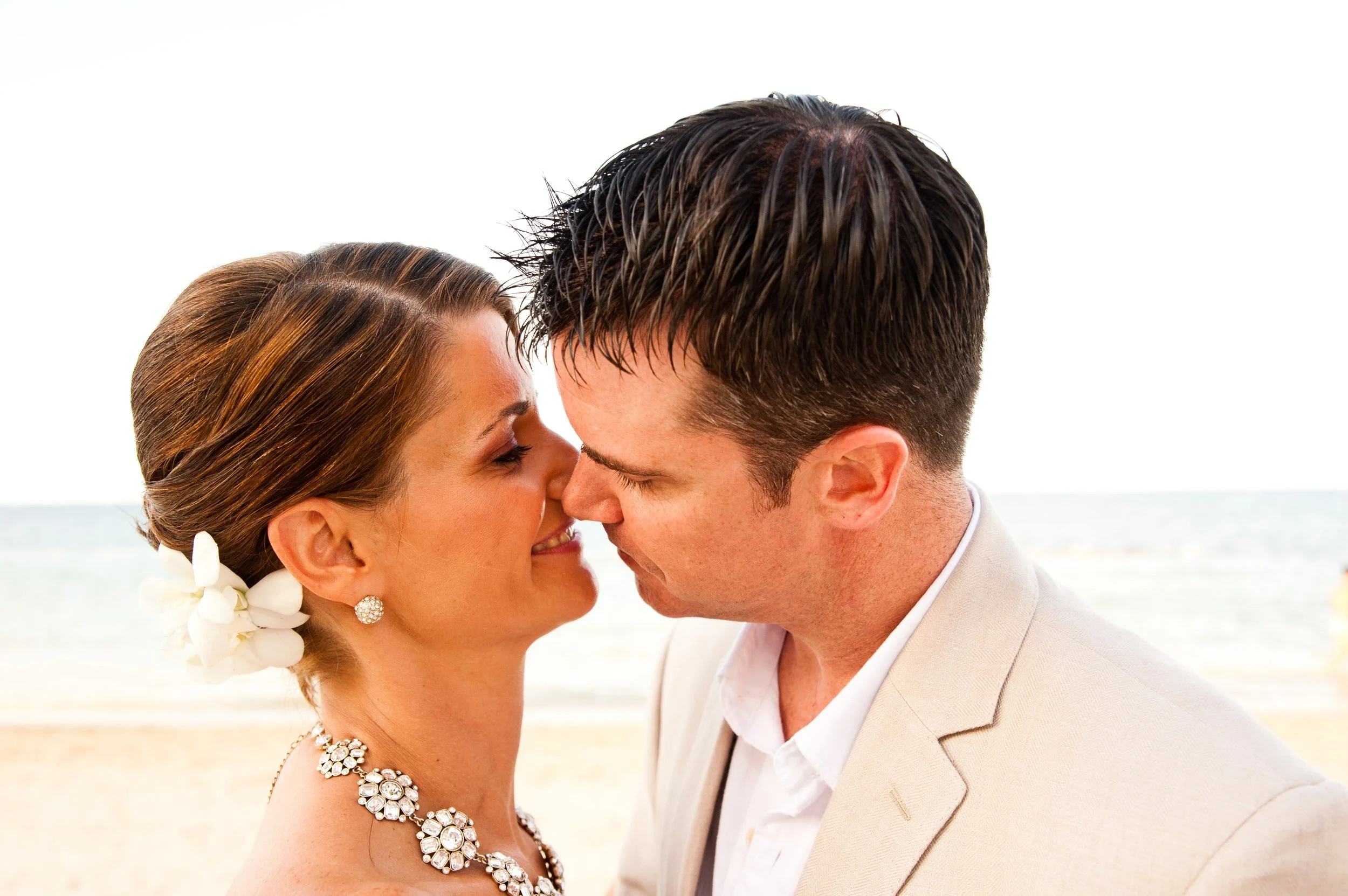 A bride and groom face each other with their foreheads and noses touching, smiling at each other on a beach with the ocean in the background.