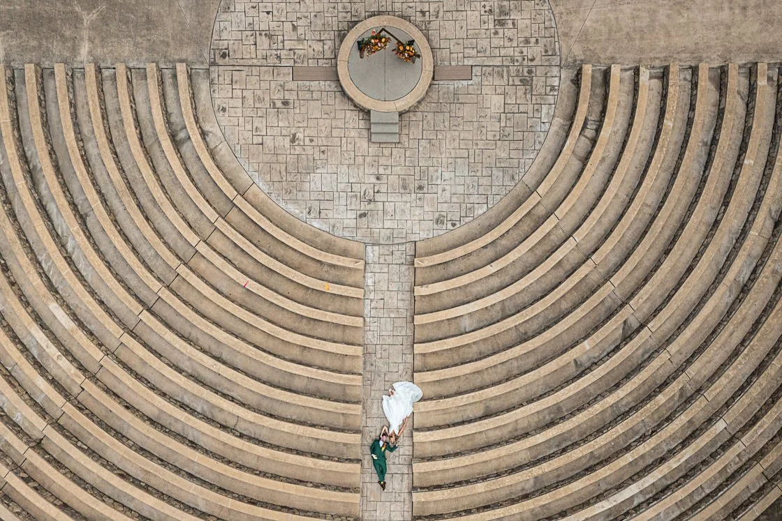 An aerial view of a wedding ceremony with a bride in a white dress and a groom in a green suit standing on a stone aisle. The ceremony is set outdoors with curved amphitheater-style seating and a small altar with some flower arrangements.