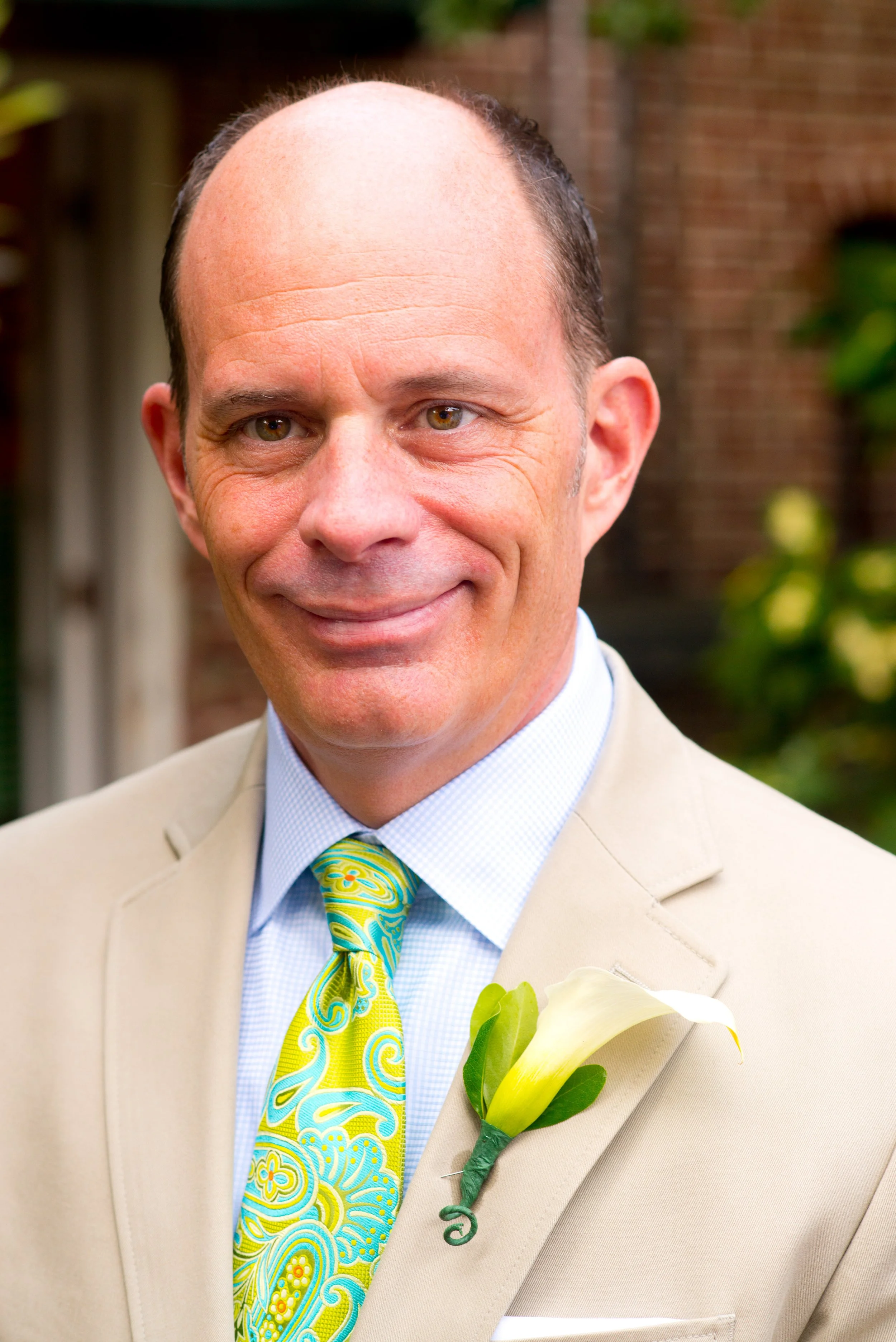A man in a beige suit with a yellow and blue paisley tie and a white collared shirt, wearing a boutonniere with white calla lily and greenery, outdoors with greenery and a brick wall in the background.