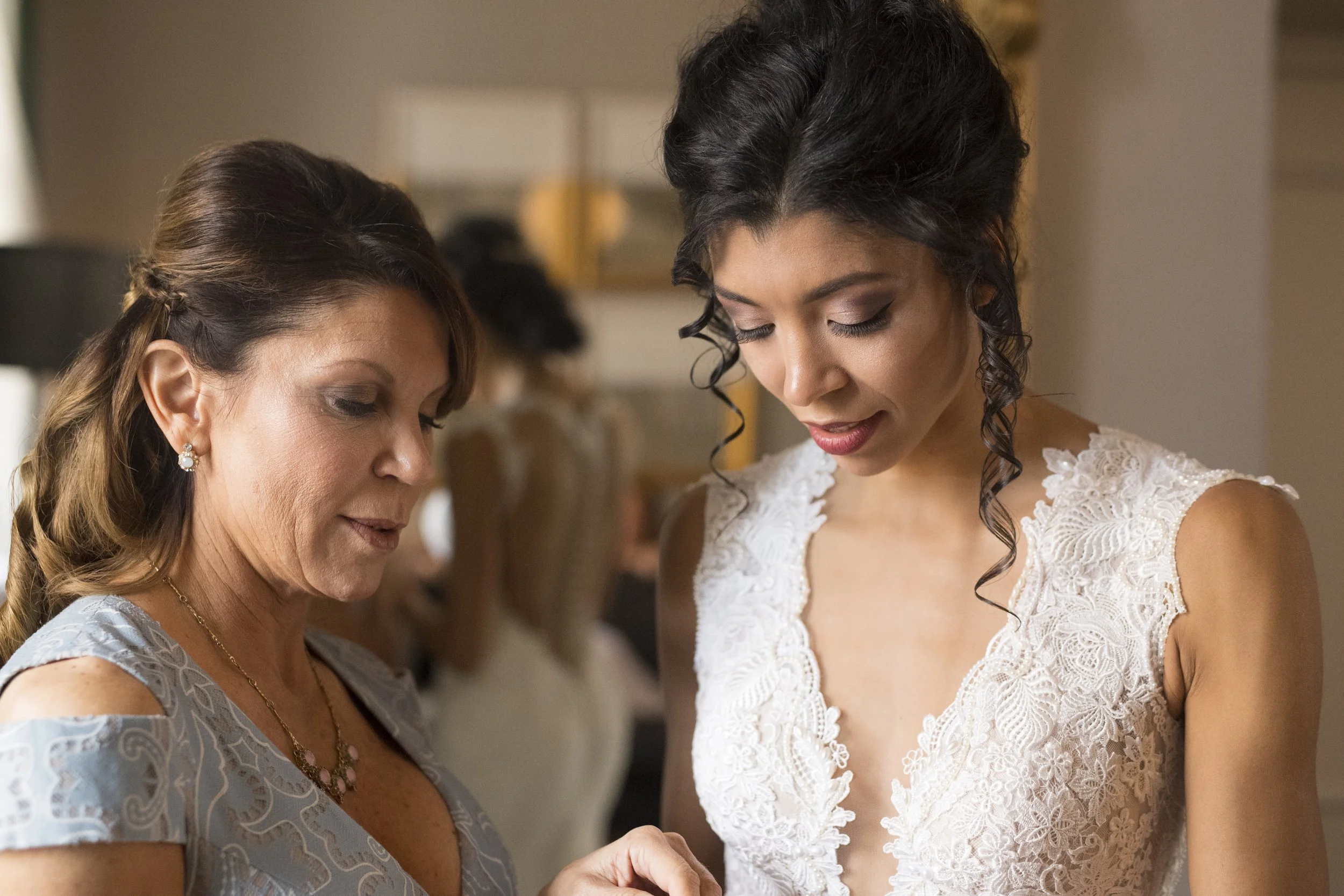 Two women looking down, one in a light blue dress, the other in a white lace dress, with a blurred background of indoor setting.