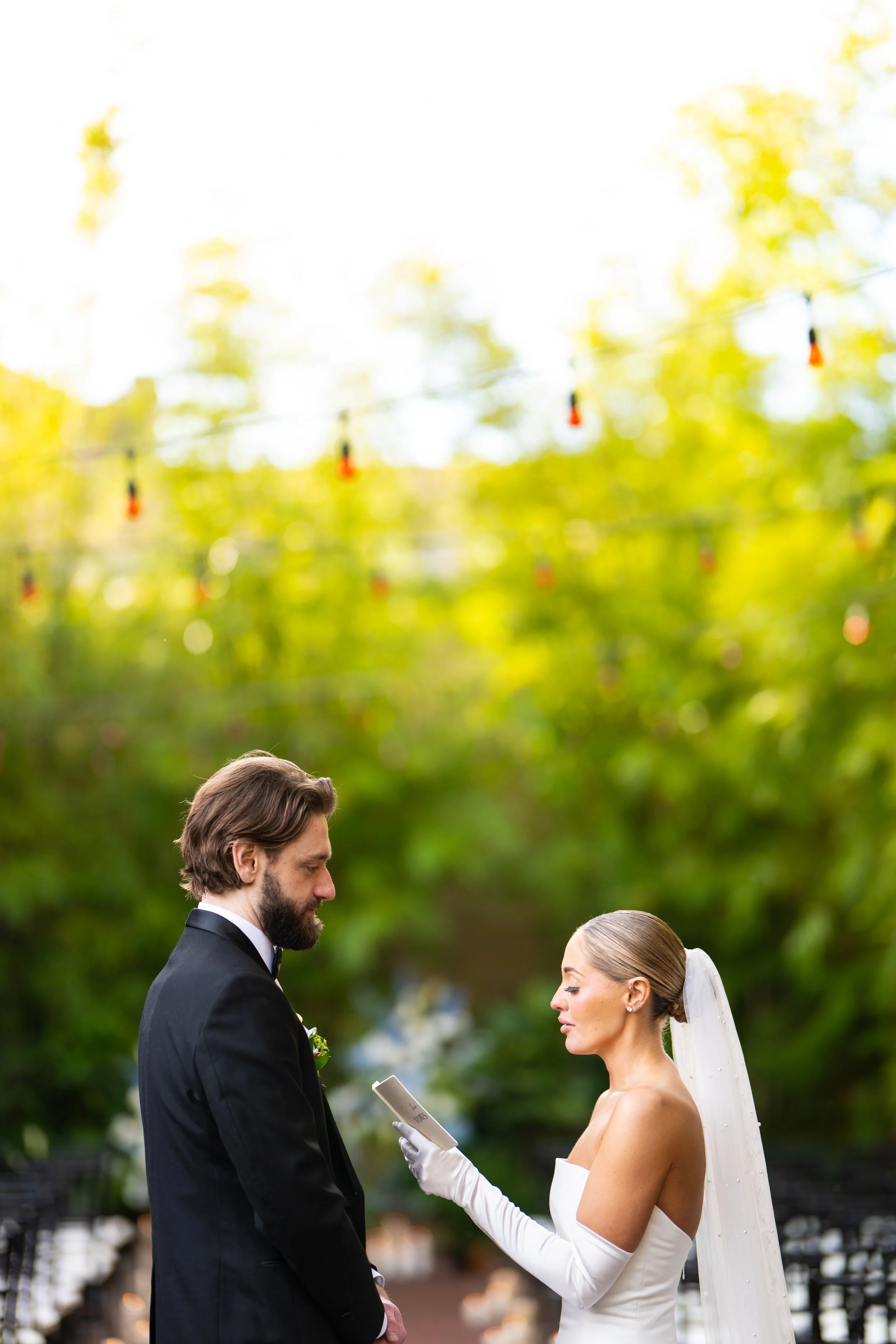 A bride and groom exchanging vows during their outdoor wedding ceremony under a canopy of trees with hanging lights.