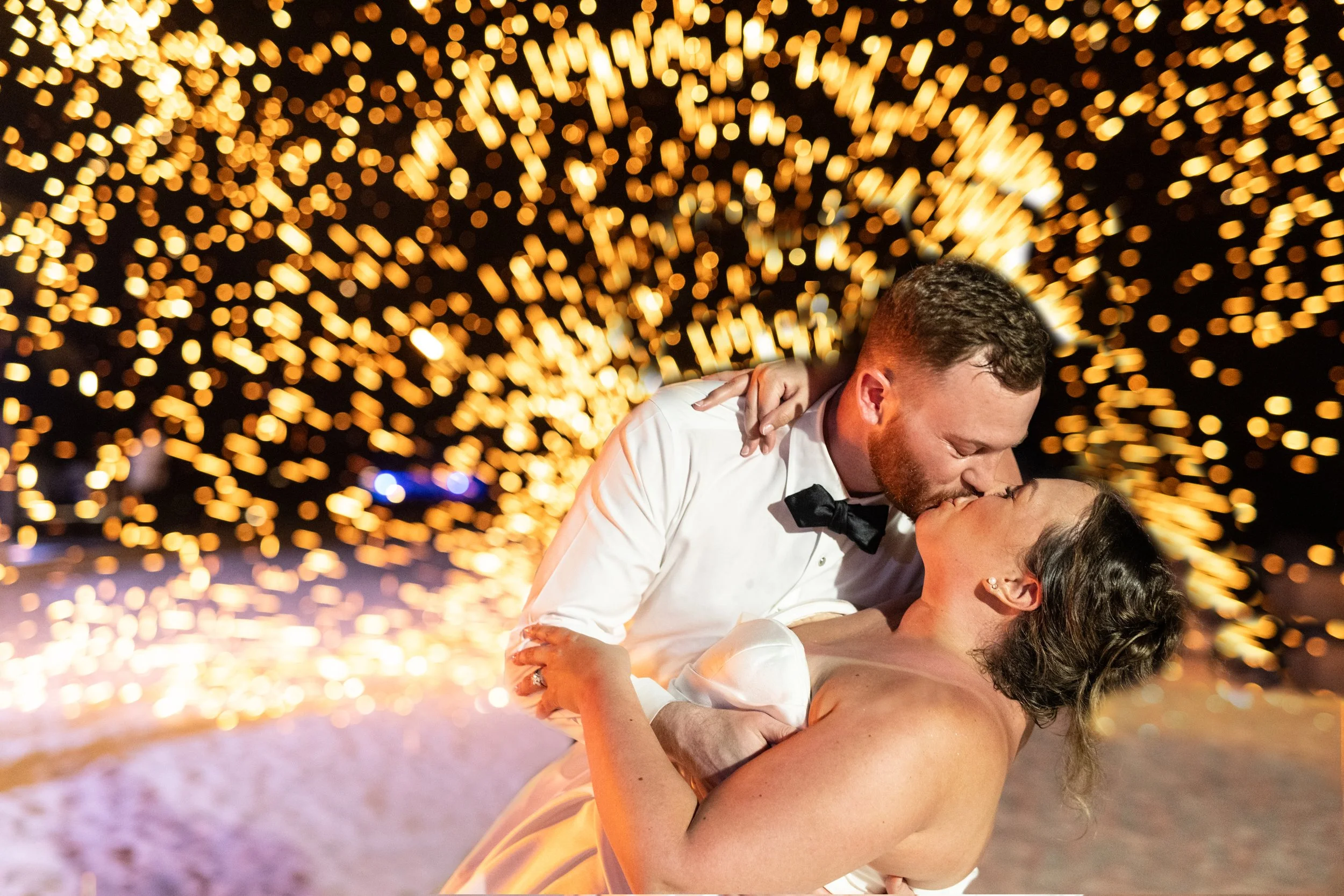 A man in a tuxedo and a woman in a dress are dancing closely on a beach at night. The background is illuminated with numerous golden bokeh lights creating a festive atmosphere.