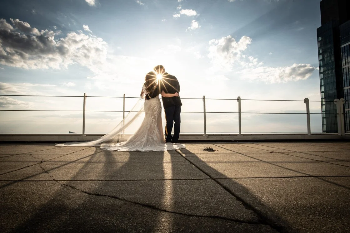 A couple sharing a kiss on a rooftop during sunset, with the sun creating a starburst effect behind them. The bride in a wedding gown and veil, and the groom in a dark suit, stand on a cracked concrete surface with a city skyline in the background.