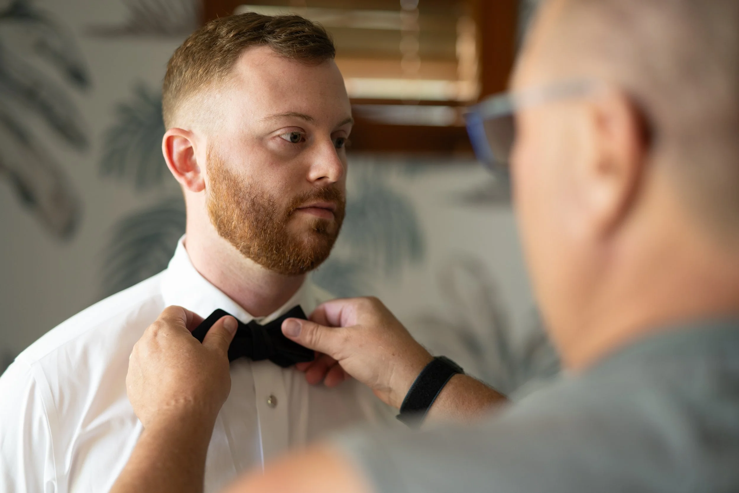 A man with red hair and a beard getting dressed in formalwear, assisted by a person adjusting a black bow tie around his neck in a room with wooden blinds.