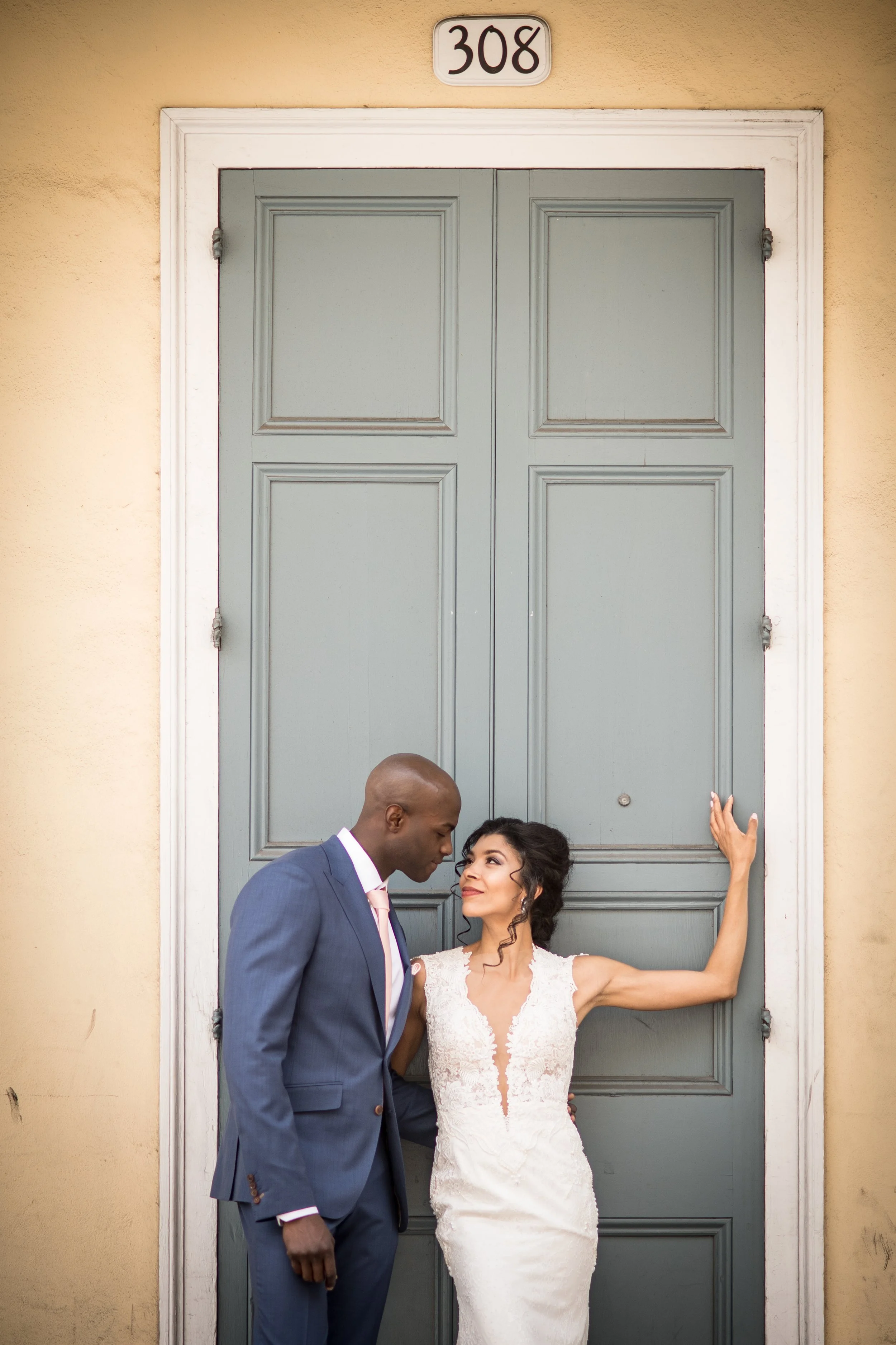 A couple, with the woman in a white lace wedding dress and the man in a blue suit, standing in front of a large green door with the number 308 above it, engaging in an intimate moment.