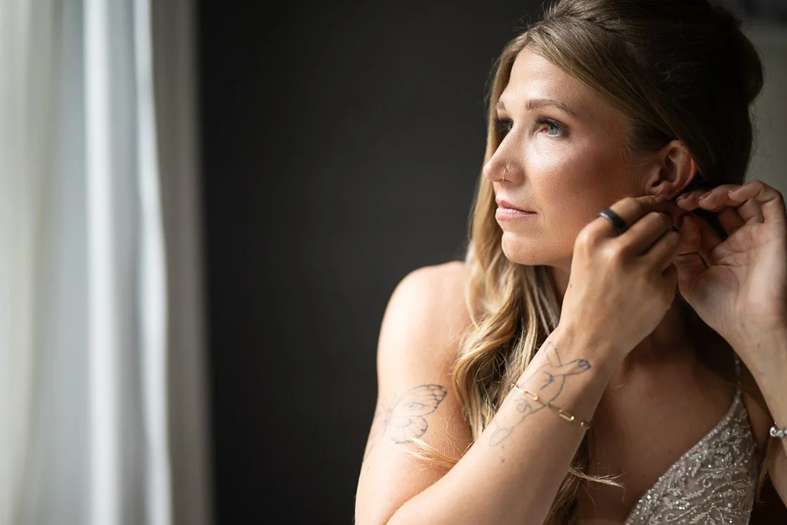 A woman with blonde hair and tattoos puts on earrings near a window with white curtains.