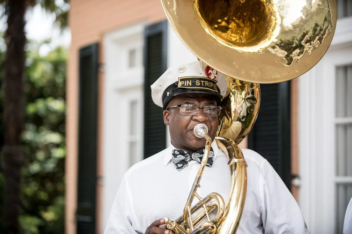 A musician playing a sousaphone outdoors, wearing a white shirt, a bow tie, glasses, and a cap with 'PIN STRIPE' on it, in front of a building with black shutters.