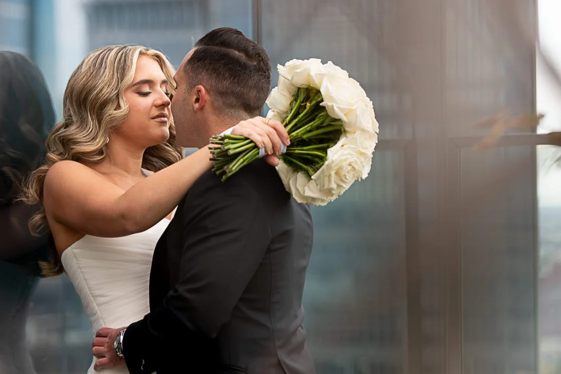 A woman in a white dress holds a bouquet of white roses, leaning in close to a man in a black suit, as if about to kiss.
