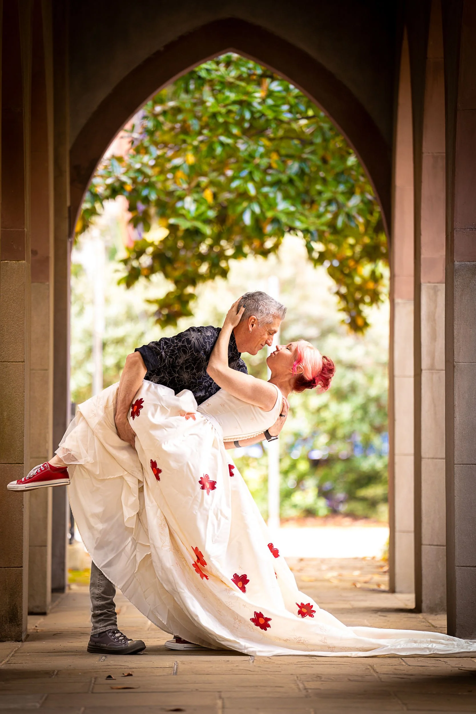A couple sharing a romantic moment, with the man lifting the woman in a white dress with red flower patterns, inside a stone archway with greenery outside.
