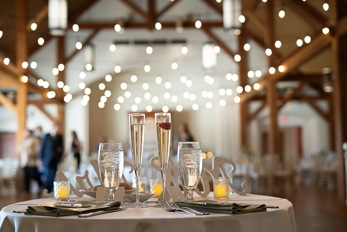 Wedding reception table with champagne glasses and water glasses, decorated with candles, in a rustic barn with string lights.