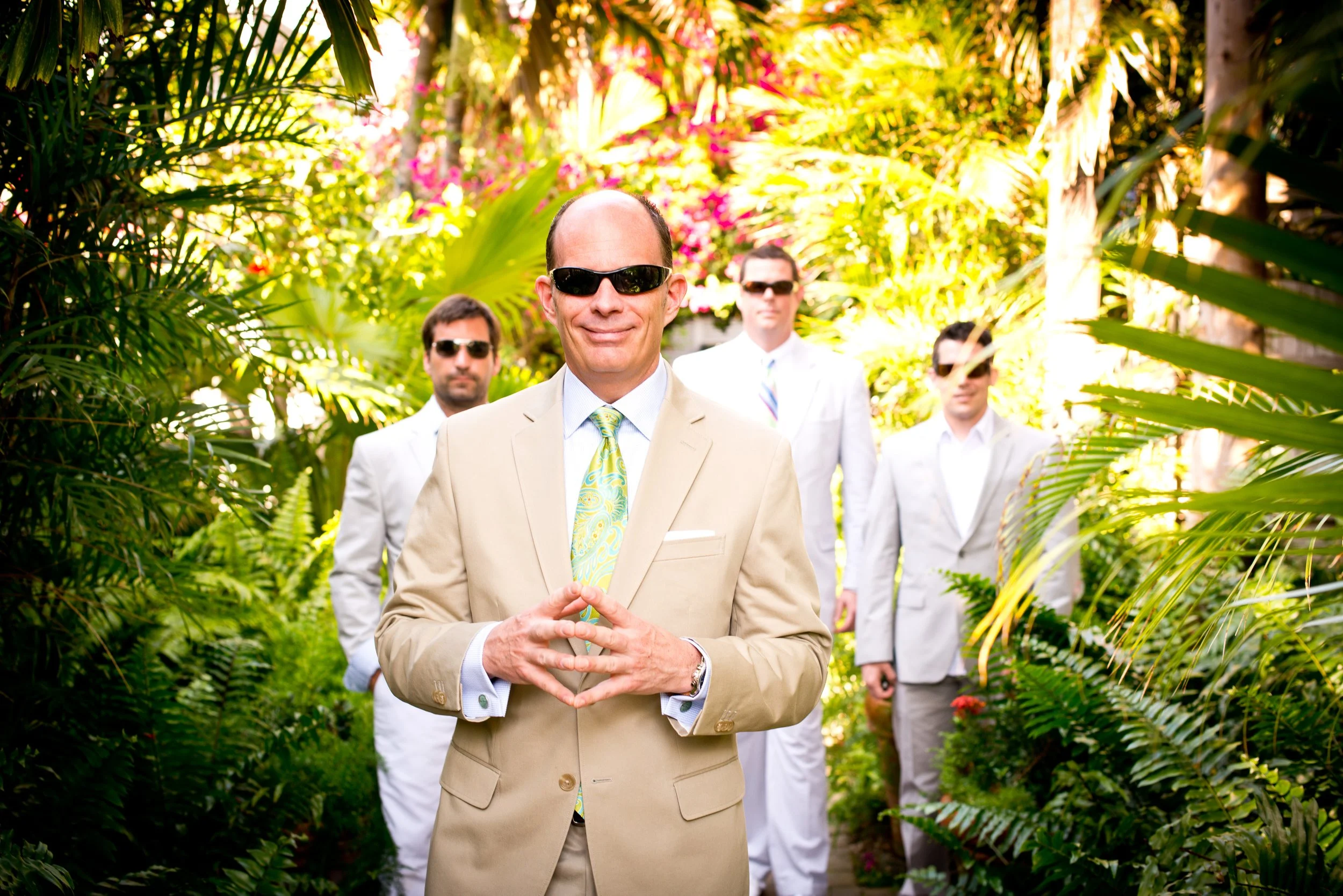 Four men in suits wearing sunglasses walking through a lush tropical garden, with the man in front smiling and clasping his hands together.
