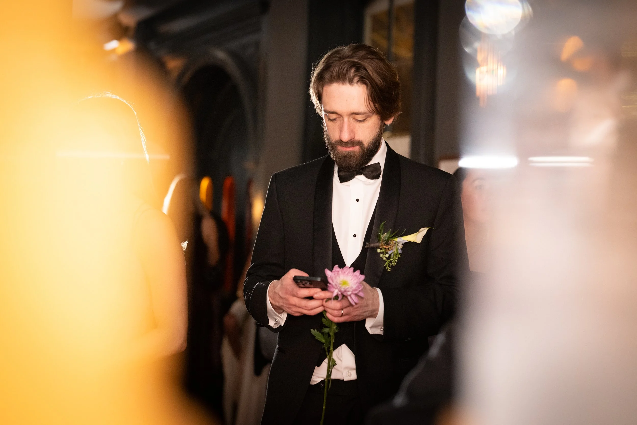 A man in a tuxedo with a bow tie and boutonniere is looking at his phone while holding a pink flower, with blurred lights in the background.