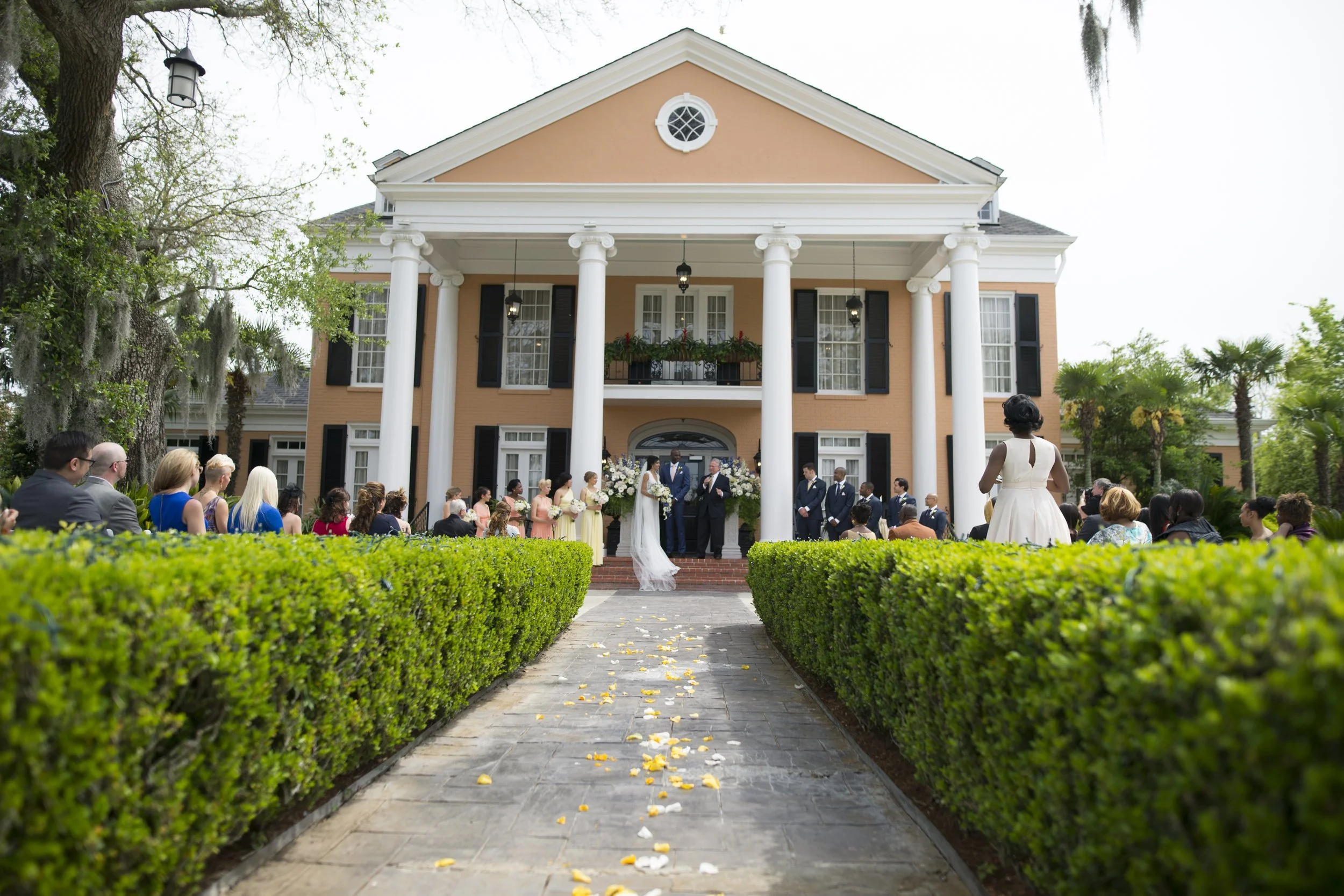 Wedding ceremony taking place outside a large peach-colored house with white columns, black shutters, and a balcony decorated with flowers. Guests seated on either side of a flower-petal-strewn aisle, watching the couple exchanging vows.