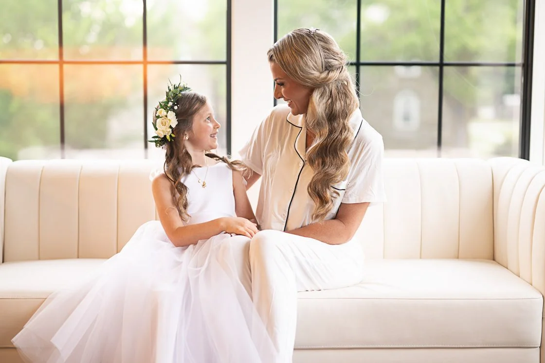 A young girl in a white dress with a floral headband sitting on a cream-colored couch, smiling and looking at an adult woman in matching pajamas, who is also smiling and looking at her, in front of large windows with a view of greenery outside.