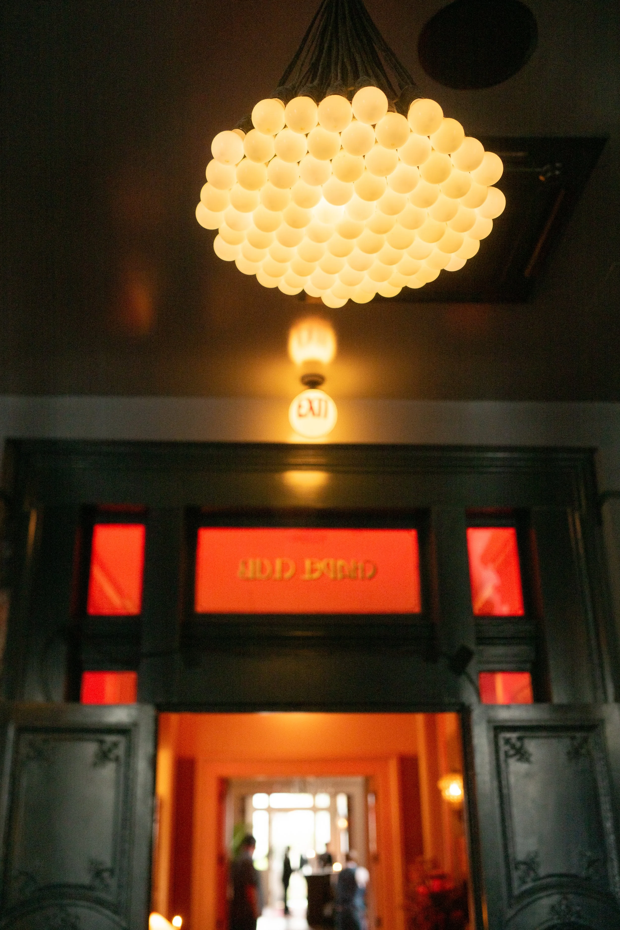 View of an ornate doorway leading into a warmly lit interior space with people and windows visible inside, framed by a dark door with decorative carvings. A chandelier with multiple spherical lights hangs from the ceiling.