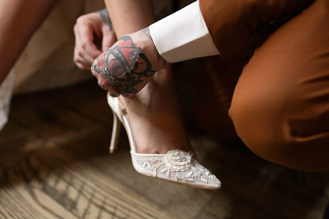Close-up of someone using a tattooed hand to help a woman put on a white high-heeled shoe with decorative embroidery.