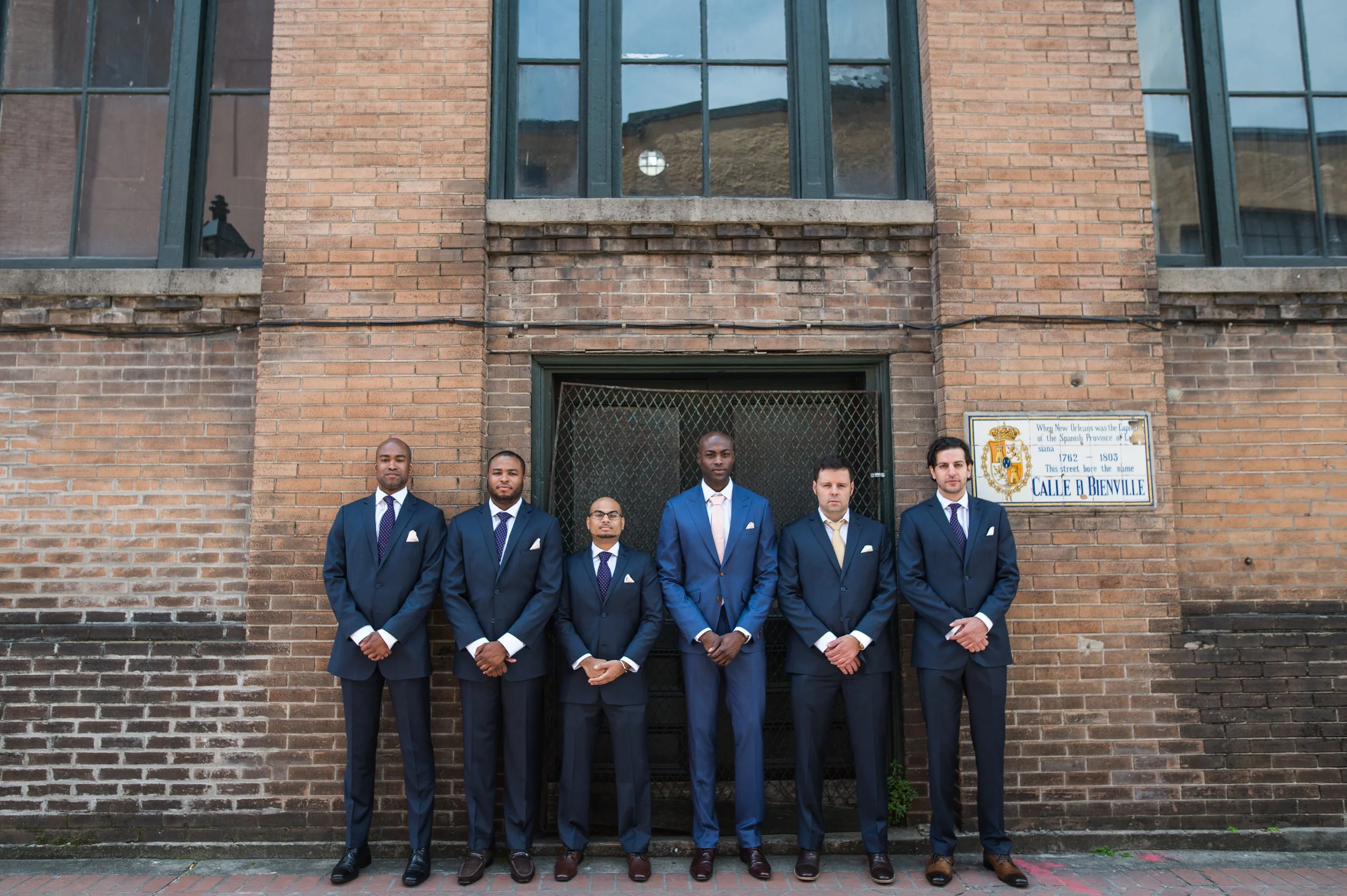 Six men dressed in formal suits standing in front of a brick building with large windows. One of the men is wearing glasses, and they are all looking at the camera.