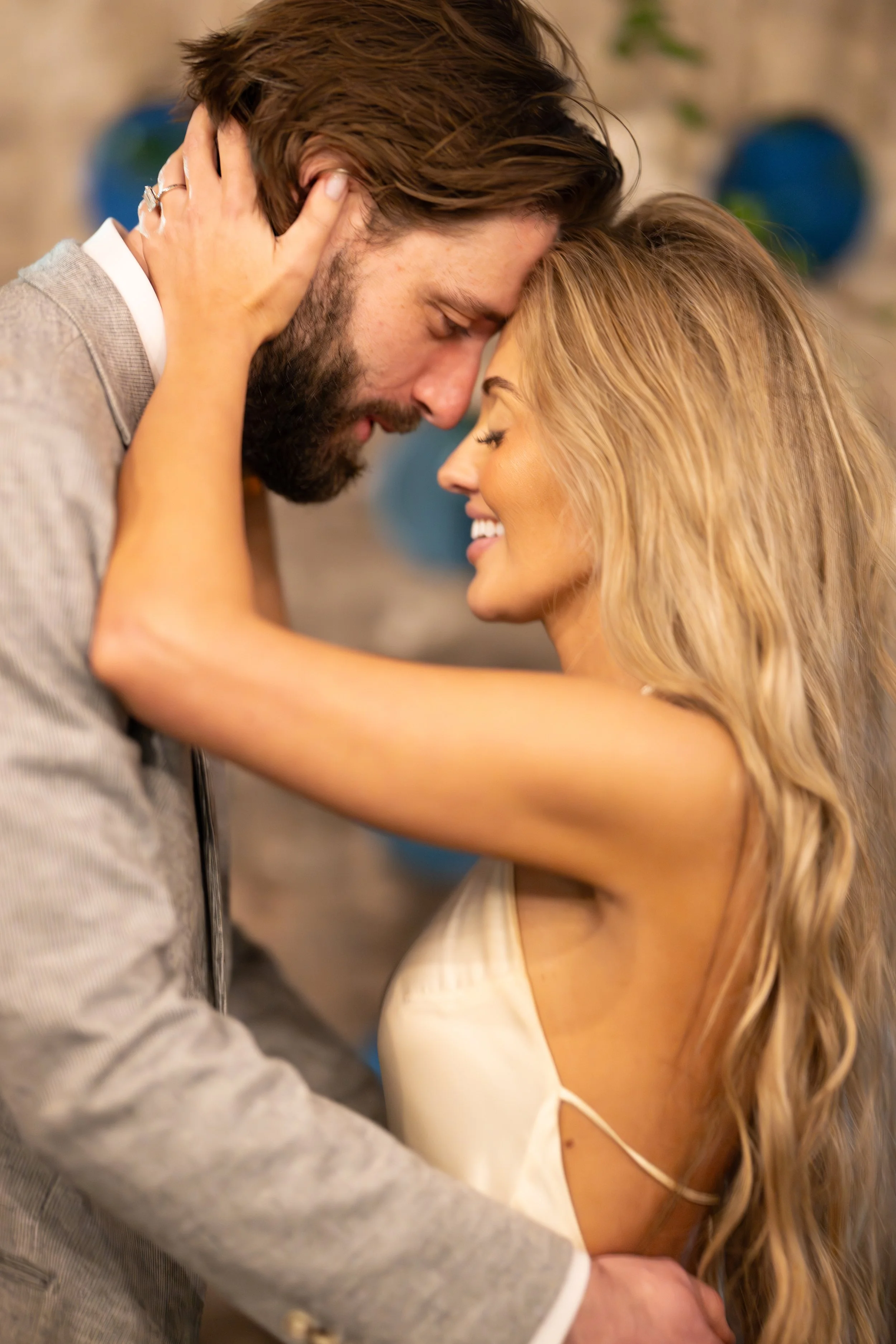 A couple sharing an intimate moment, about to kiss, with their foreheads touching and eyes closed, smiling gently. The man has brown hair and a beard, and the woman has long, blonde hair. They are in a warm, softly-lit indoor setting.