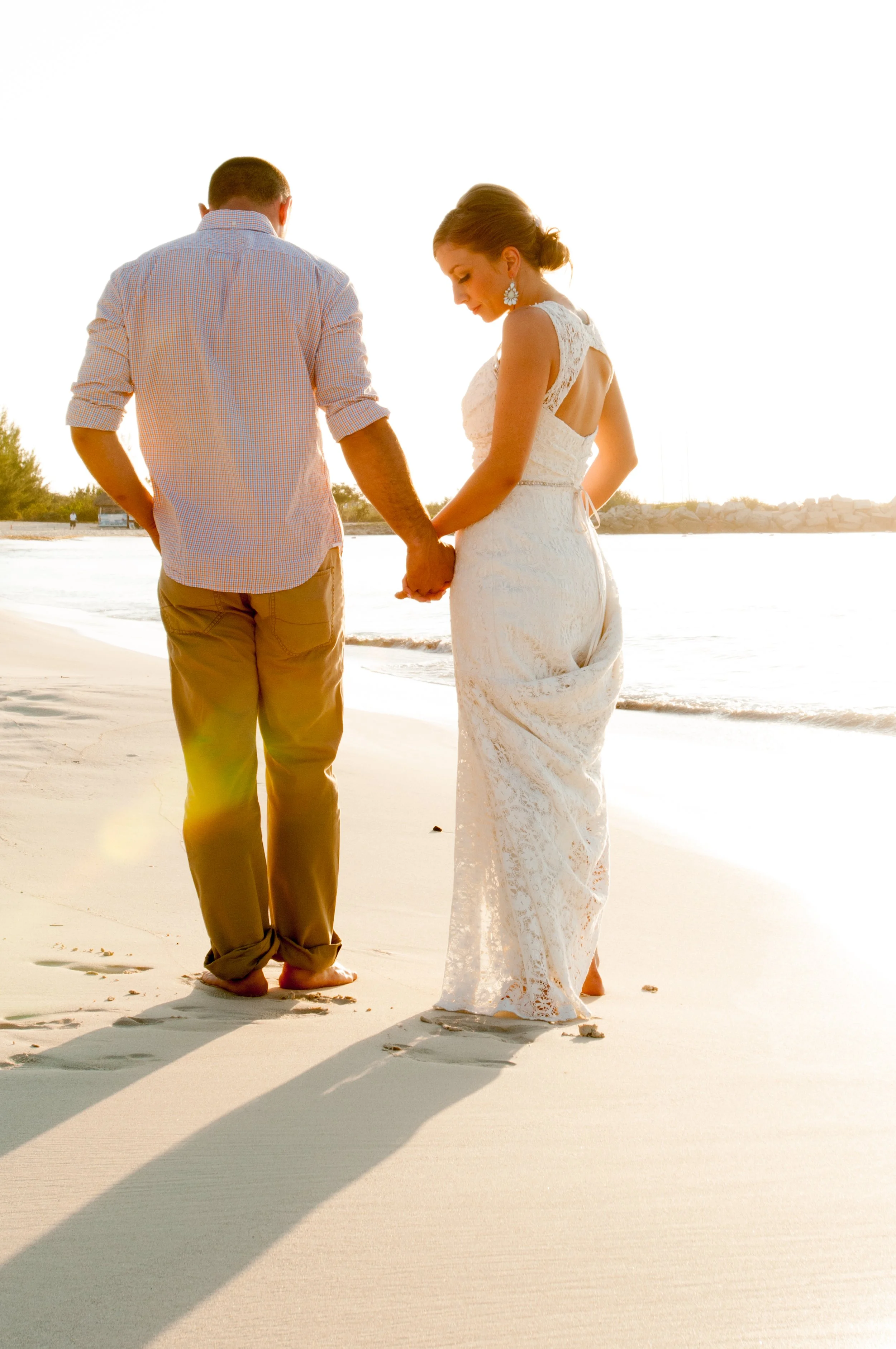 A couple holding hands on a beach at sunset, with the woman in a white lace dress and the man in a light button-up shirt and khakis, both barefoot, standing near the shoreline.