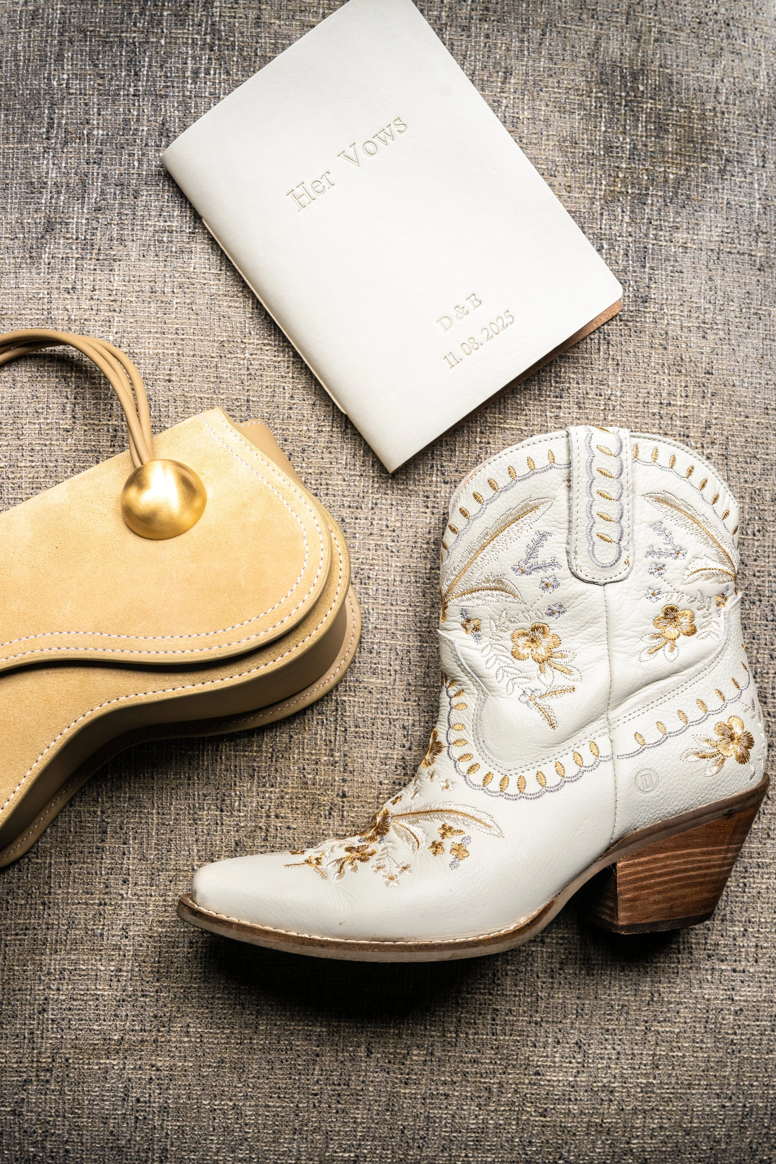 A white cowboy boot with floral embroidery, a beige handbag with a rounded handle, and a white book with embossed gold lettering on a textured beige surface.