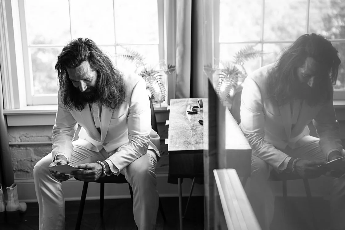 A man with long hair and a beard, dressed in a suit, sitting on a chair by a window, looking at his phone.