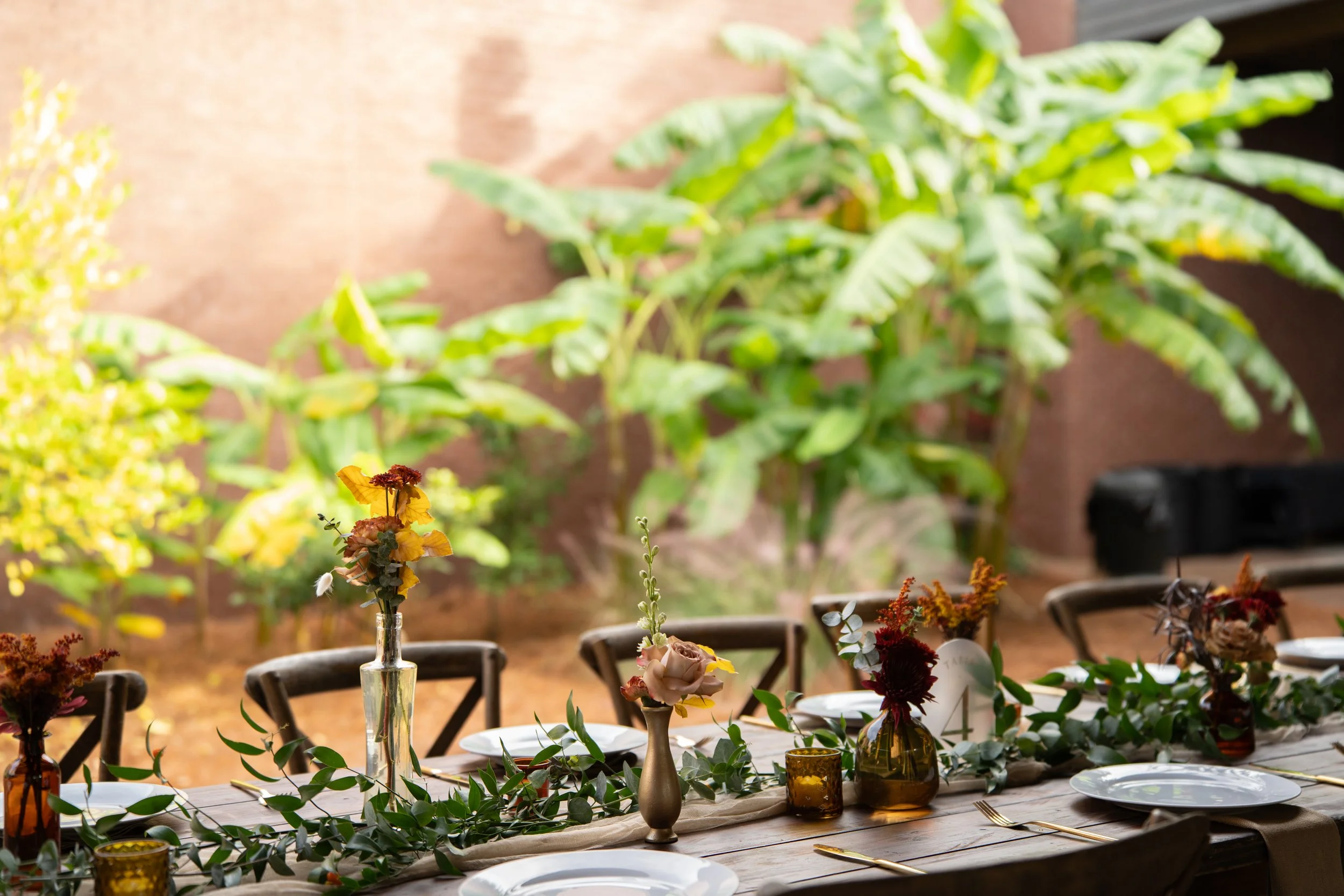 Tables set for a meal with floral arrangements and greenery, outdoors with a wooden background.