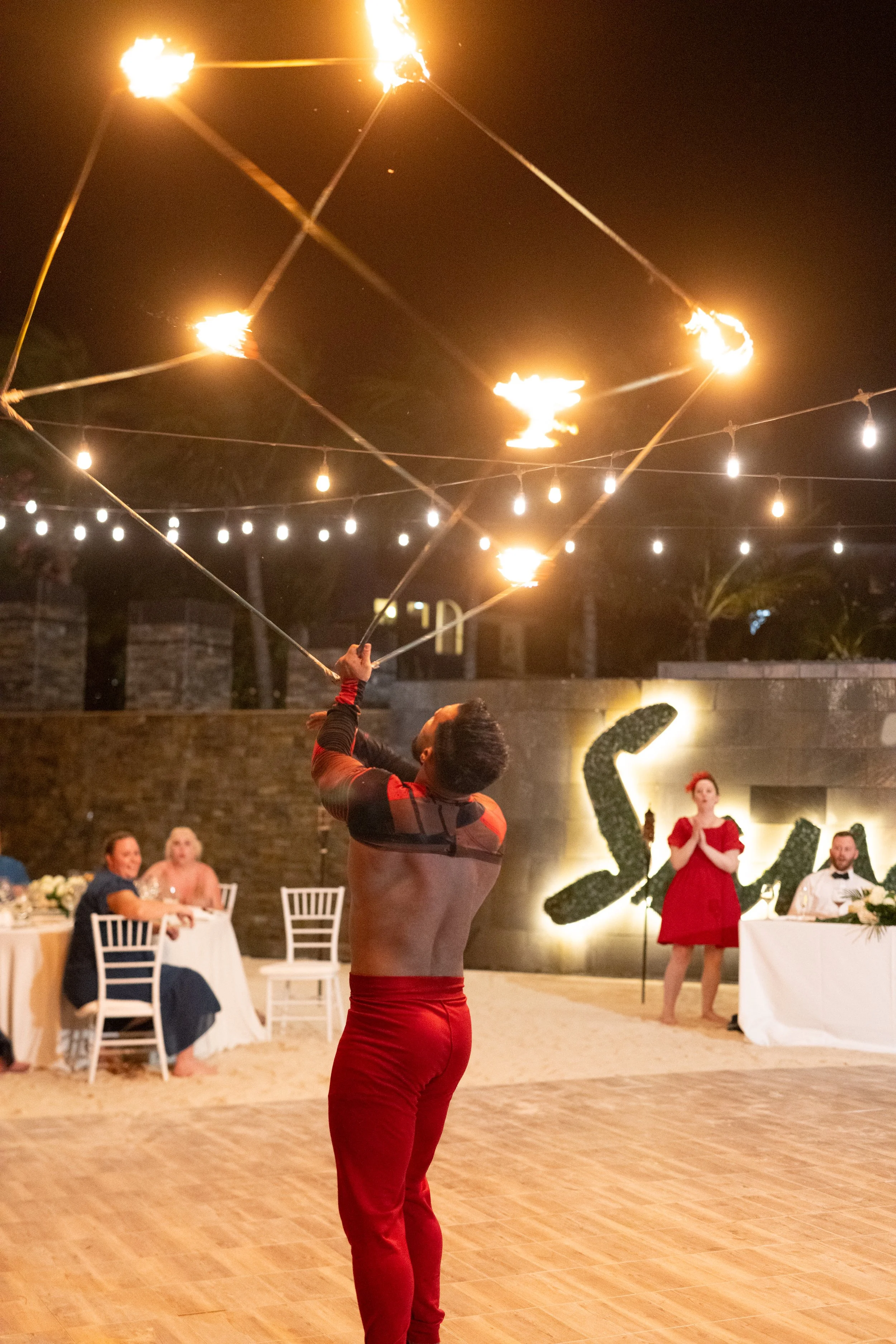 A performer spins fire poi at an outdoor event during nighttime, with guests seated at tables watching and string lights hanging overhead.