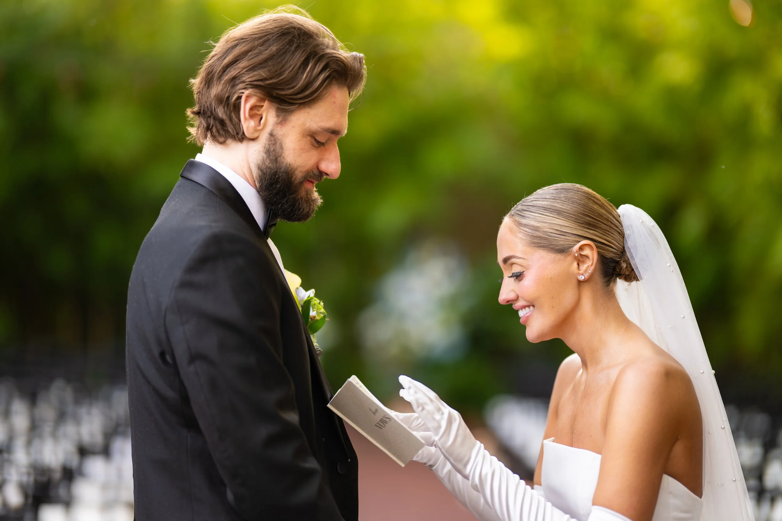 Bride and groom exchanging vows at outdoor wedding ceremony, with bride smiling and holding a book, groom in tuxedo with boutonniere, blurred green trees background.
