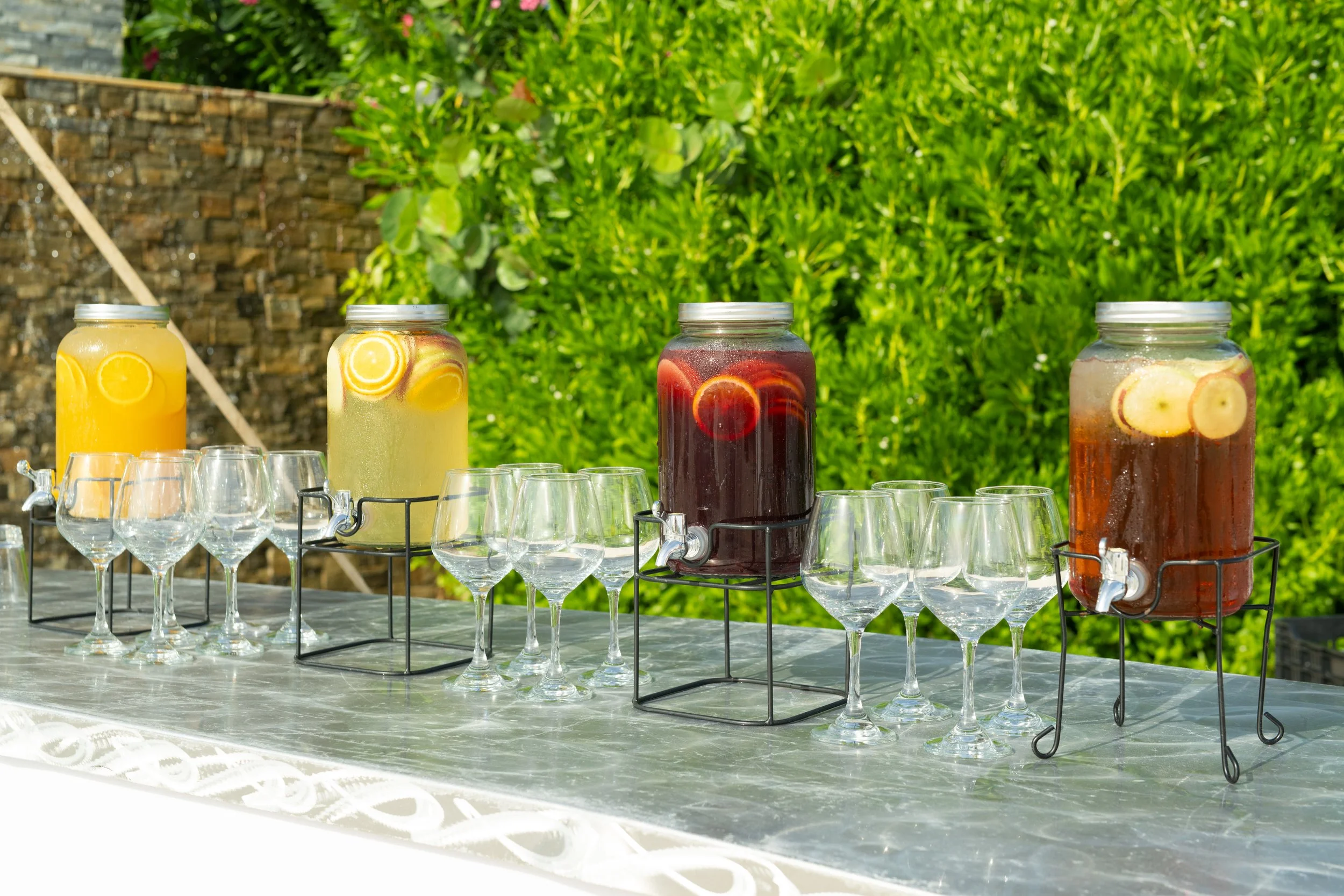 Four large glass beverage dispensers filled with colorful infused drinks, placed on black metal stands on a table outdoors, with empty wine glasses lined up in front. Lush green bushes and a stone wall are visible in the background.