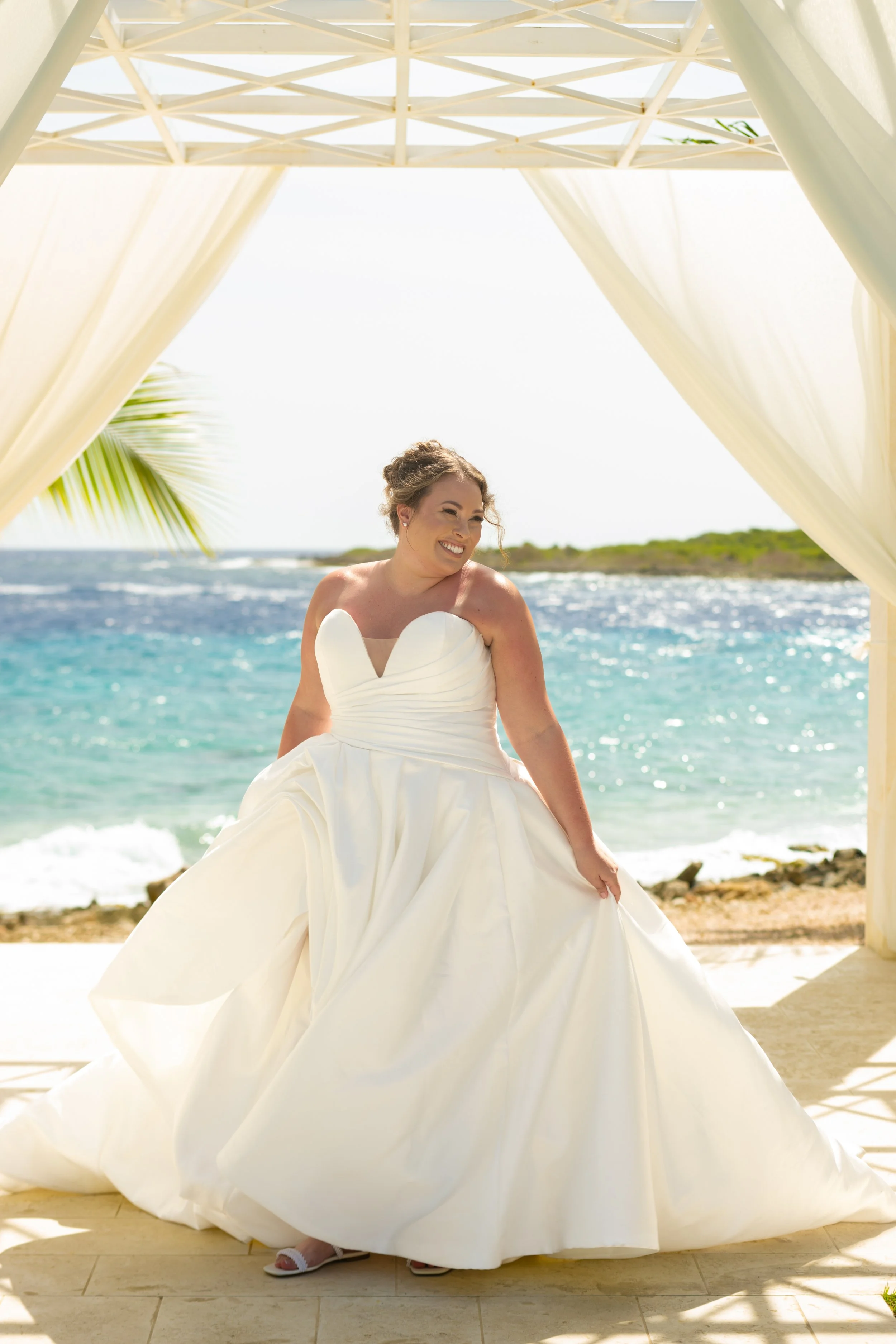 Woman in a white wedding dress standing outdoors under a canopy on the beach, with ocean and greenery in the background, smiling.