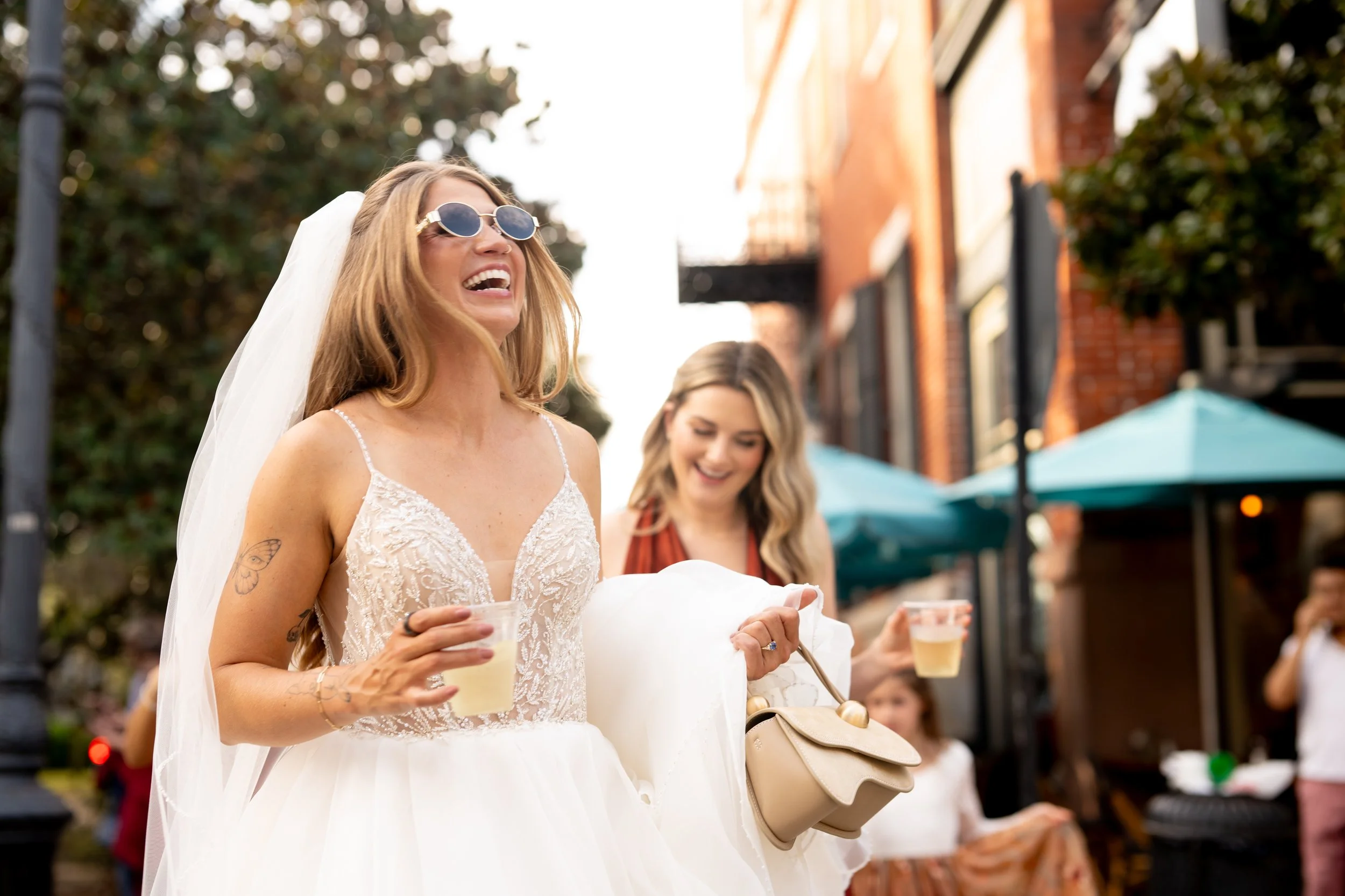 A woman in a wedding dress wearing sunglasses and smiling, holding a drink, standing outdoors at a gathering or celebration.