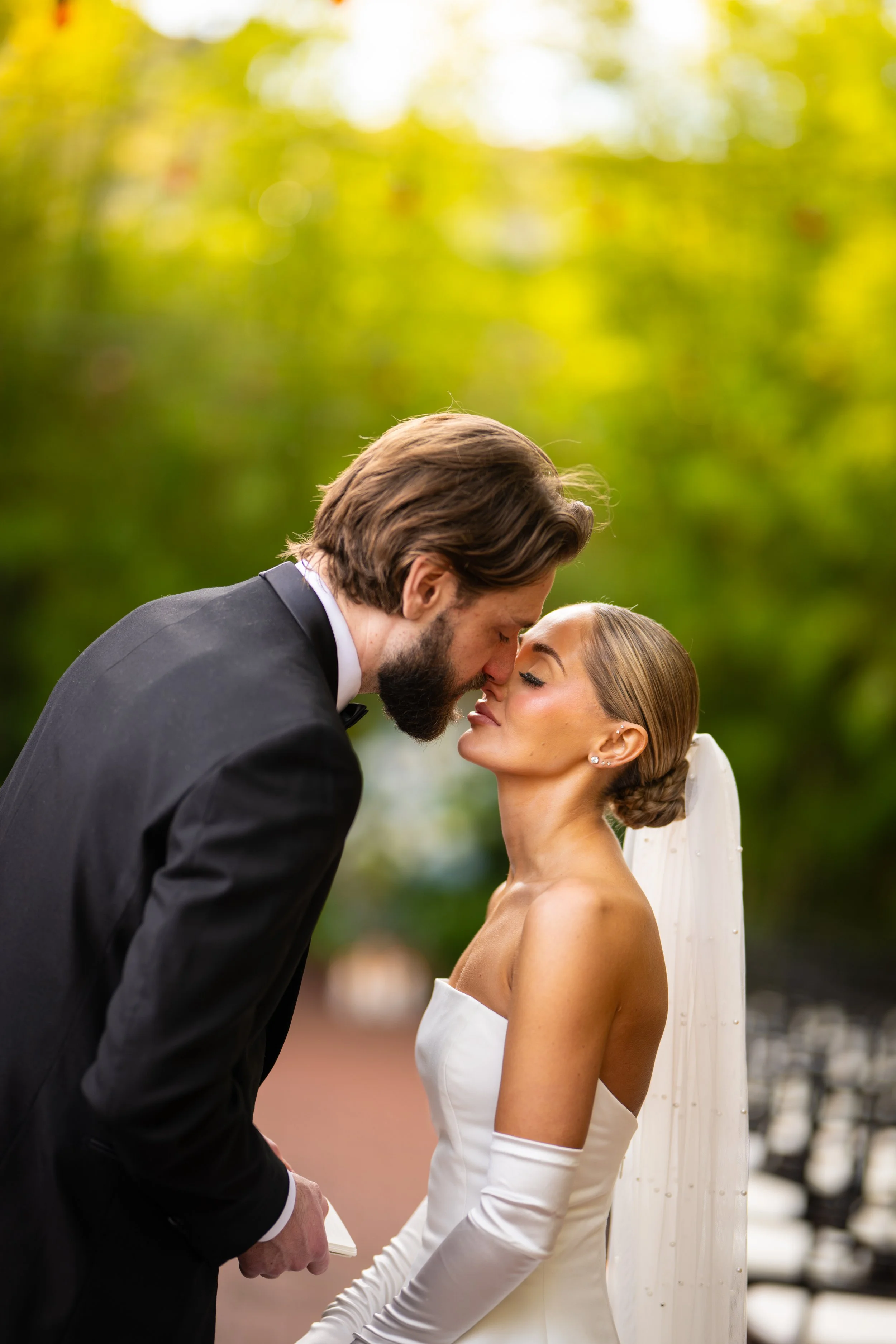 A bride and groom exchange vows outdoors, with the groom leaning forward and the bride with eyes closed in a moment of intimacy, surrounded by blurred trees and sunlight.