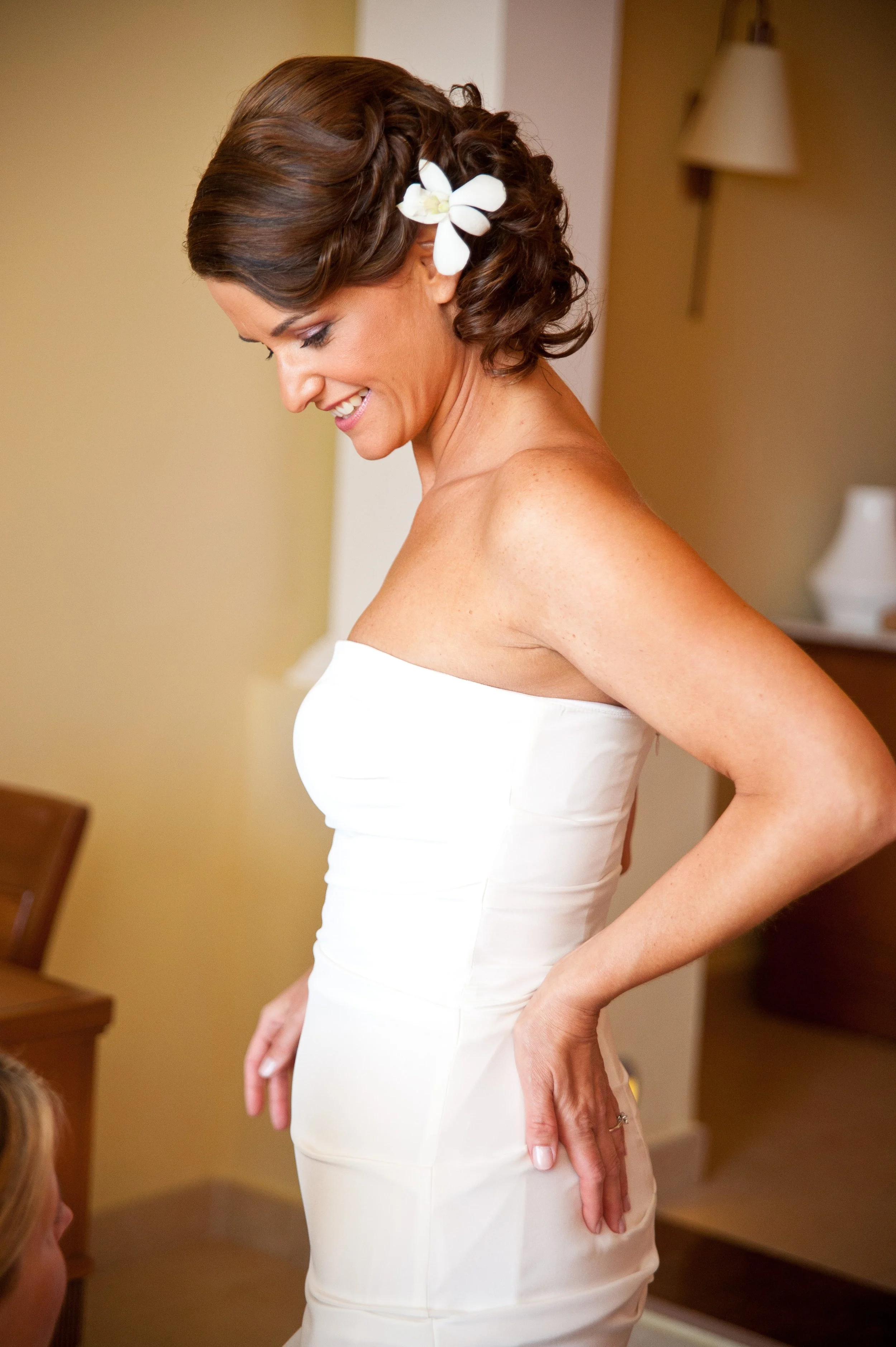 A woman with short brown hair styled in waves, wearing a white strapless dress and a white flower in her hair, smiling and looking down.