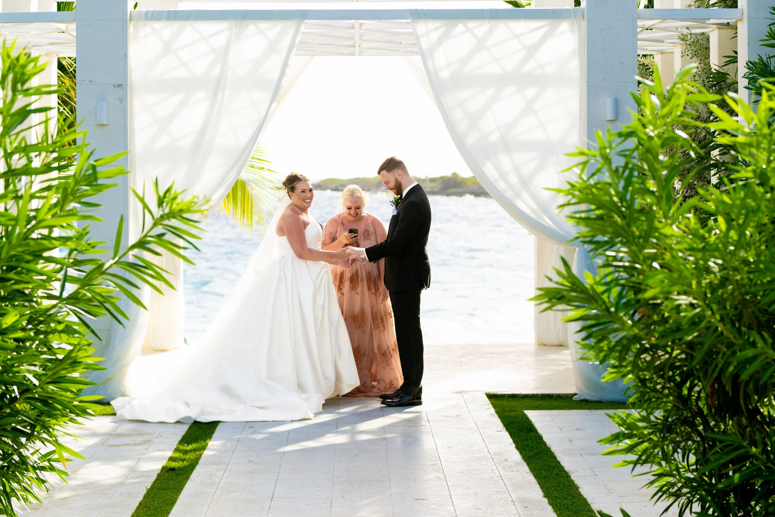 A bride and groom holding hands and smiling during a wedding ceremony outdoors by the water, with a female officiant or witness, surrounded by green plants and white drapes.