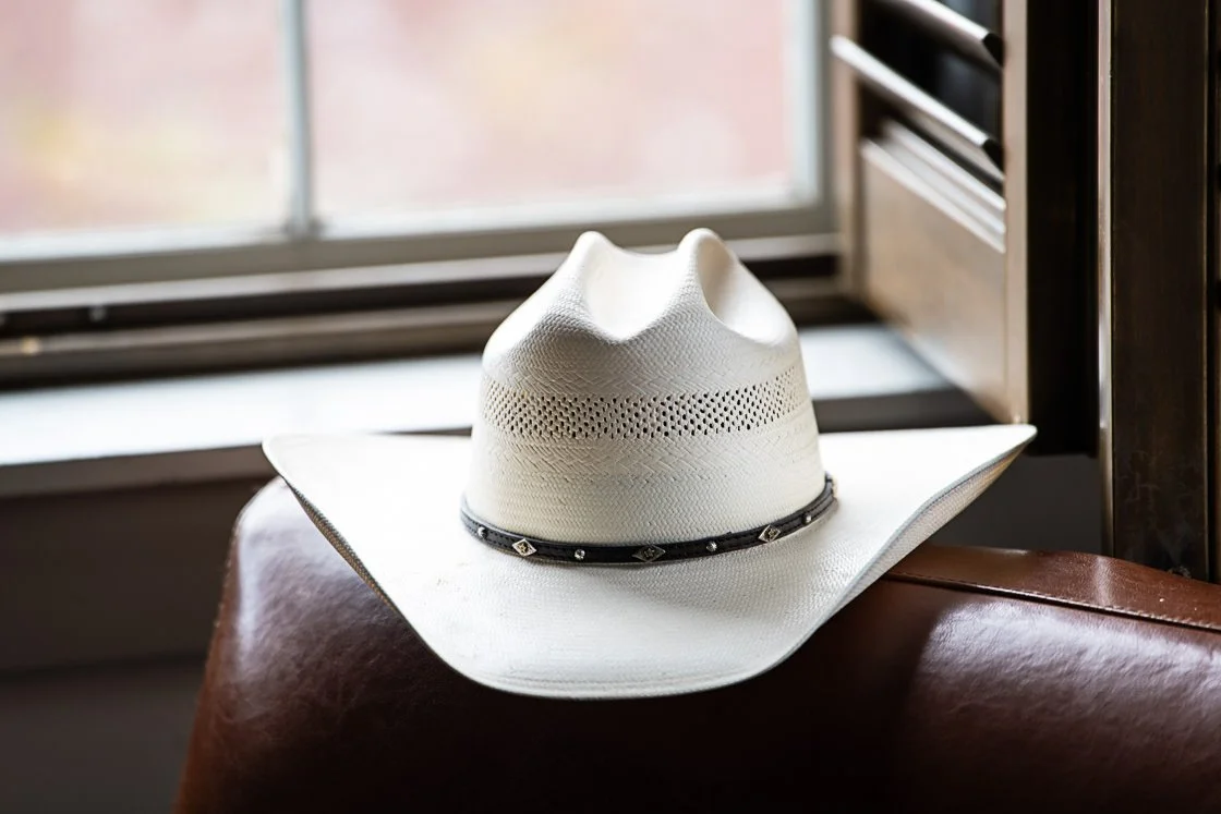 A white cowboy hat with a black band sitting on a leather chair in front of a window.