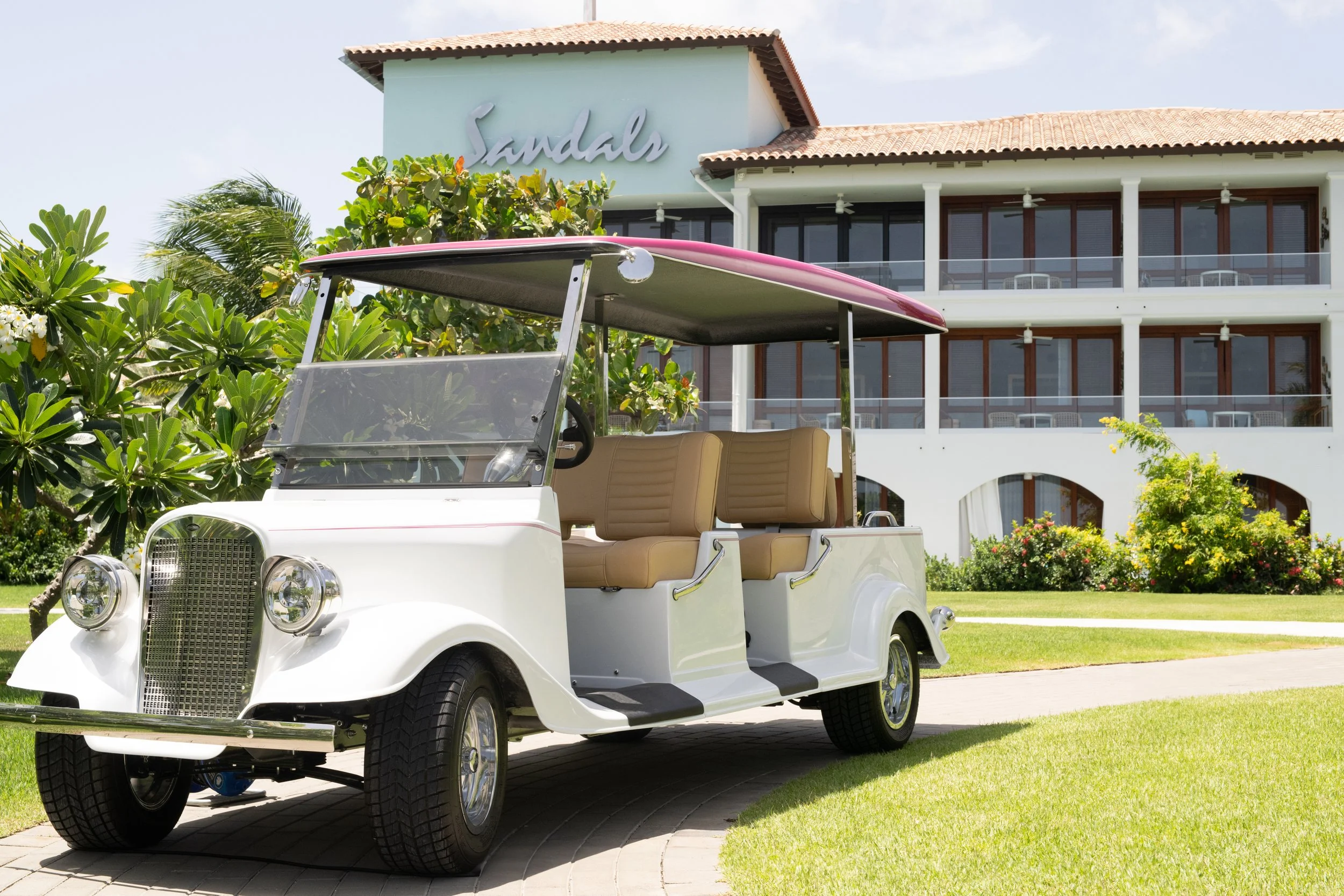 A white golf cart with beige seats parked on a brick path in front of a garden with green trees and shrubs, with a white multi-story building with a red-tiled roof and a sign that says 'Sandals' in the background.