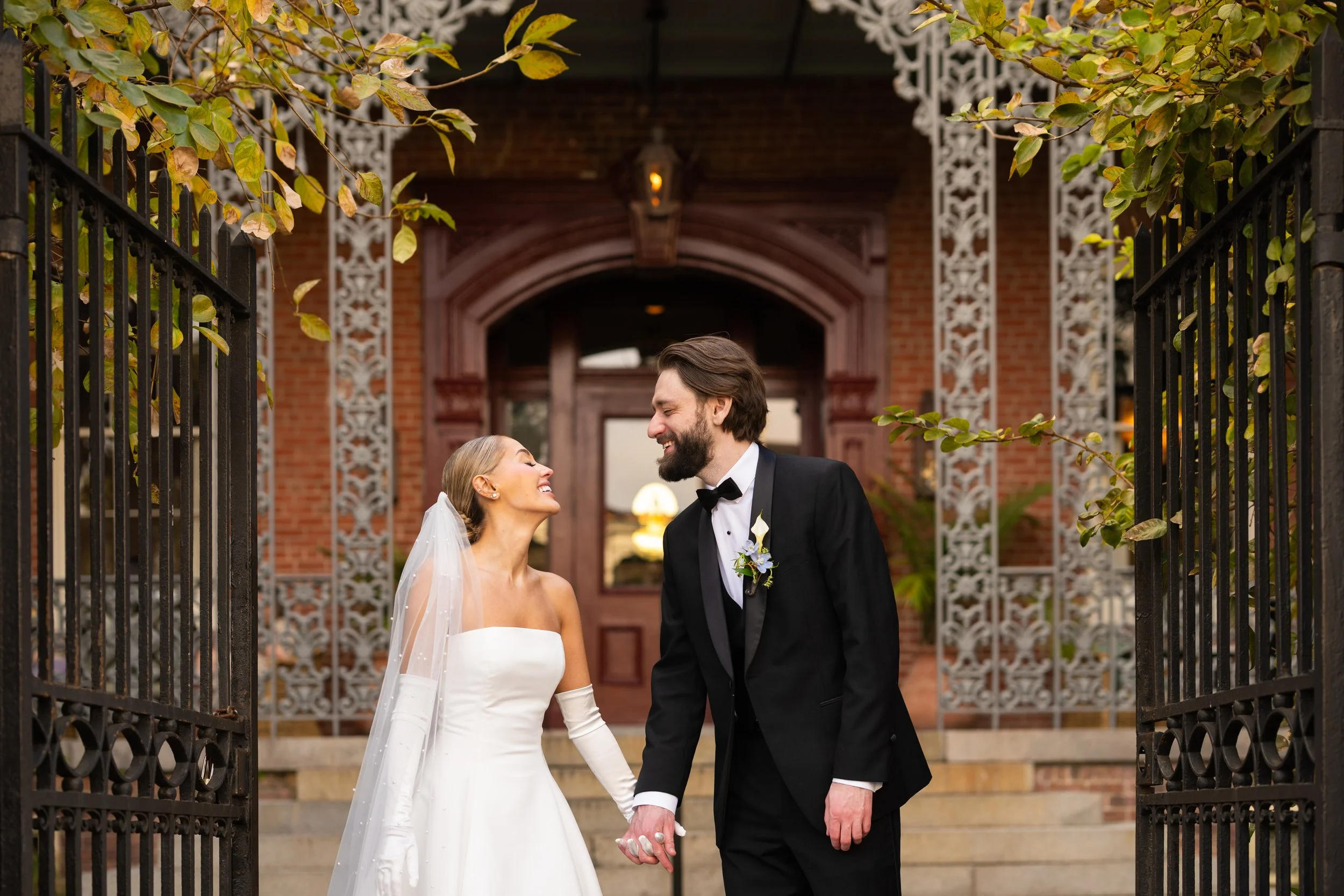 A bride and groom holding hands, smiling at each other, standing in front of an ornate brick building with decorative white trim, surrounded by open black wrought iron gates, during their wedding celebration.