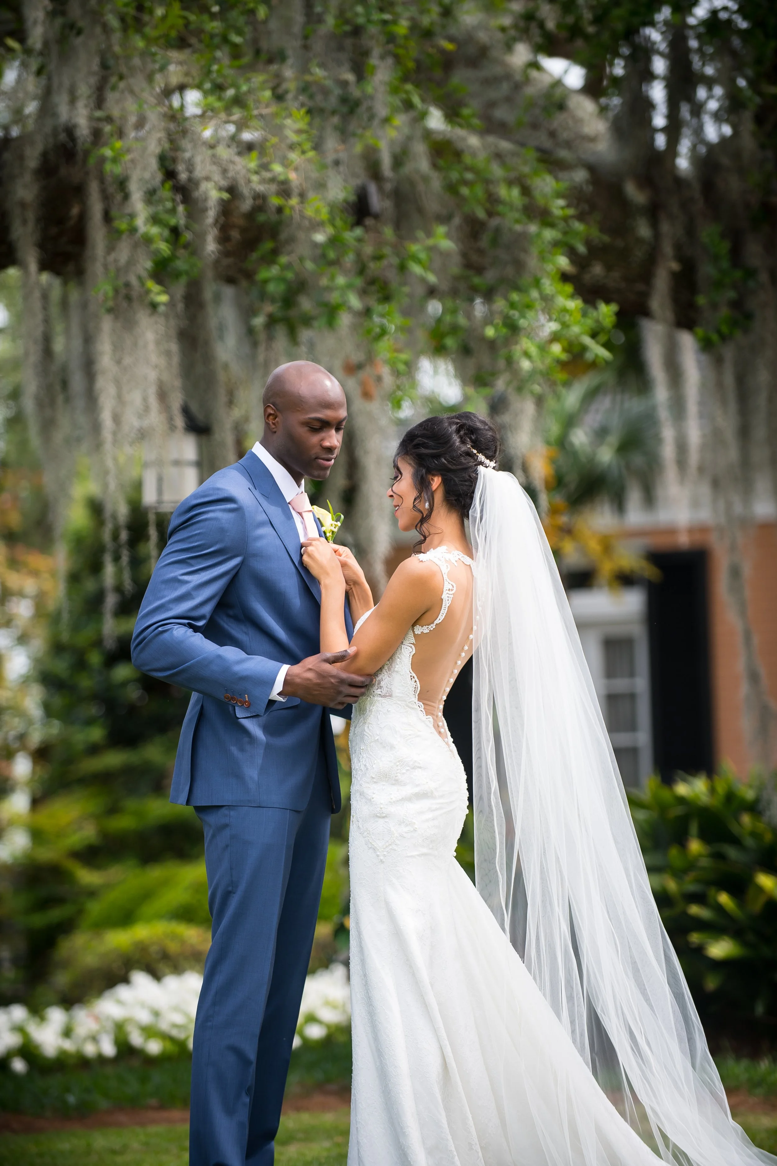 A bride and groom standing outdoors under a large tree with Spanish moss, holding hands and looking at each other during their wedding ceremony.